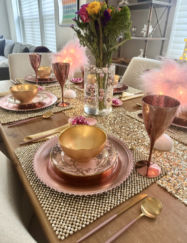 Decorated dining table with gold and pink dishes, gold cups, pink champagne glasses, floral centerpiece with pink, purple, and yellow flowers, pink feather accents, and glittery table runner.