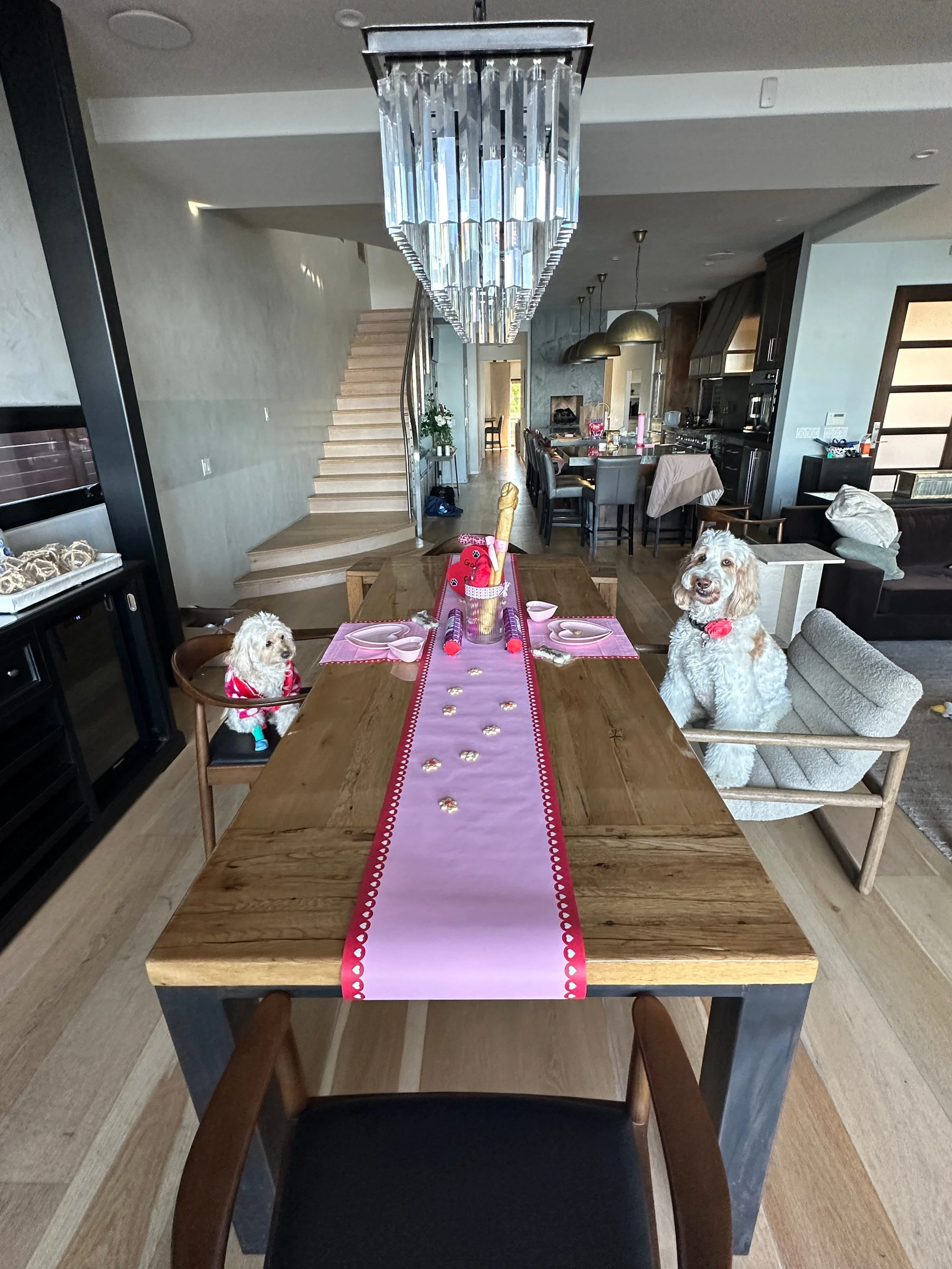 A dining table with pink heart cutout table runner, pink and white napkins, and a centerpiece with Valentine-themed decorations. Two dogs, one on a chair and one on a couch, are near the table.