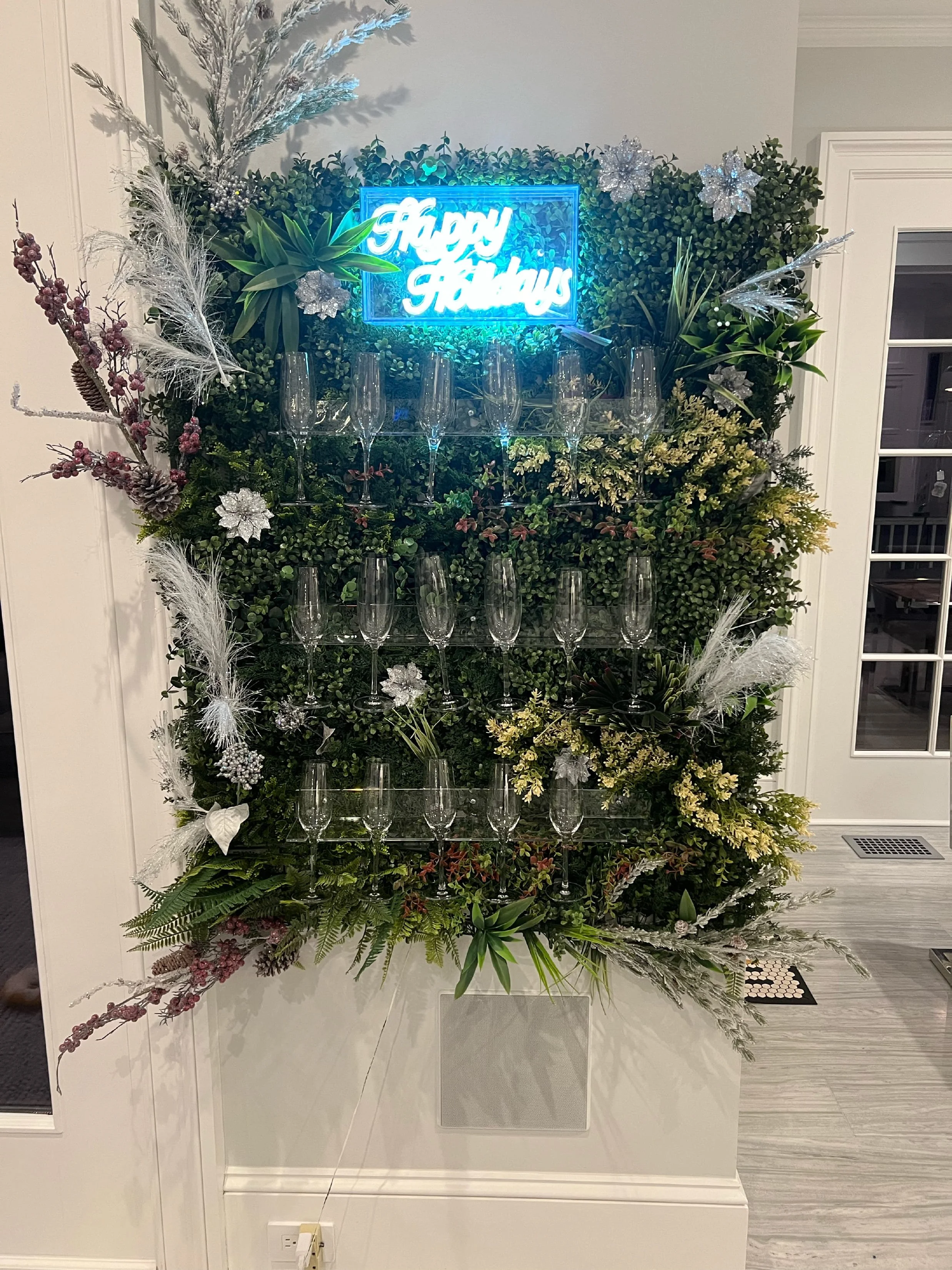 A festive holiday display featuring lush green foliage, silver and white decorative snowflakes, and an illuminated blue neon sign that reads 'Happy Holidays.' Champagne flutes are arranged on a glass shelf within the display.