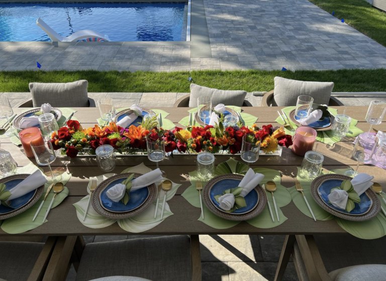 Outdoor dining table set for a group with colorful flower centerpiece, plates, glasses, napkins, and utensils near a swimming pool.