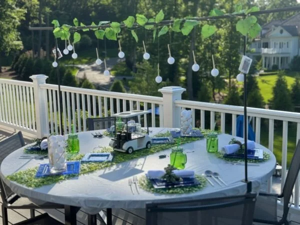 Outdoor dining table set for a celebration with a white tablecloth, green and blue glasses, plates, silverware, and napkins, decorated with string lights hanging above, on a balcony with a view of trees and houses.
