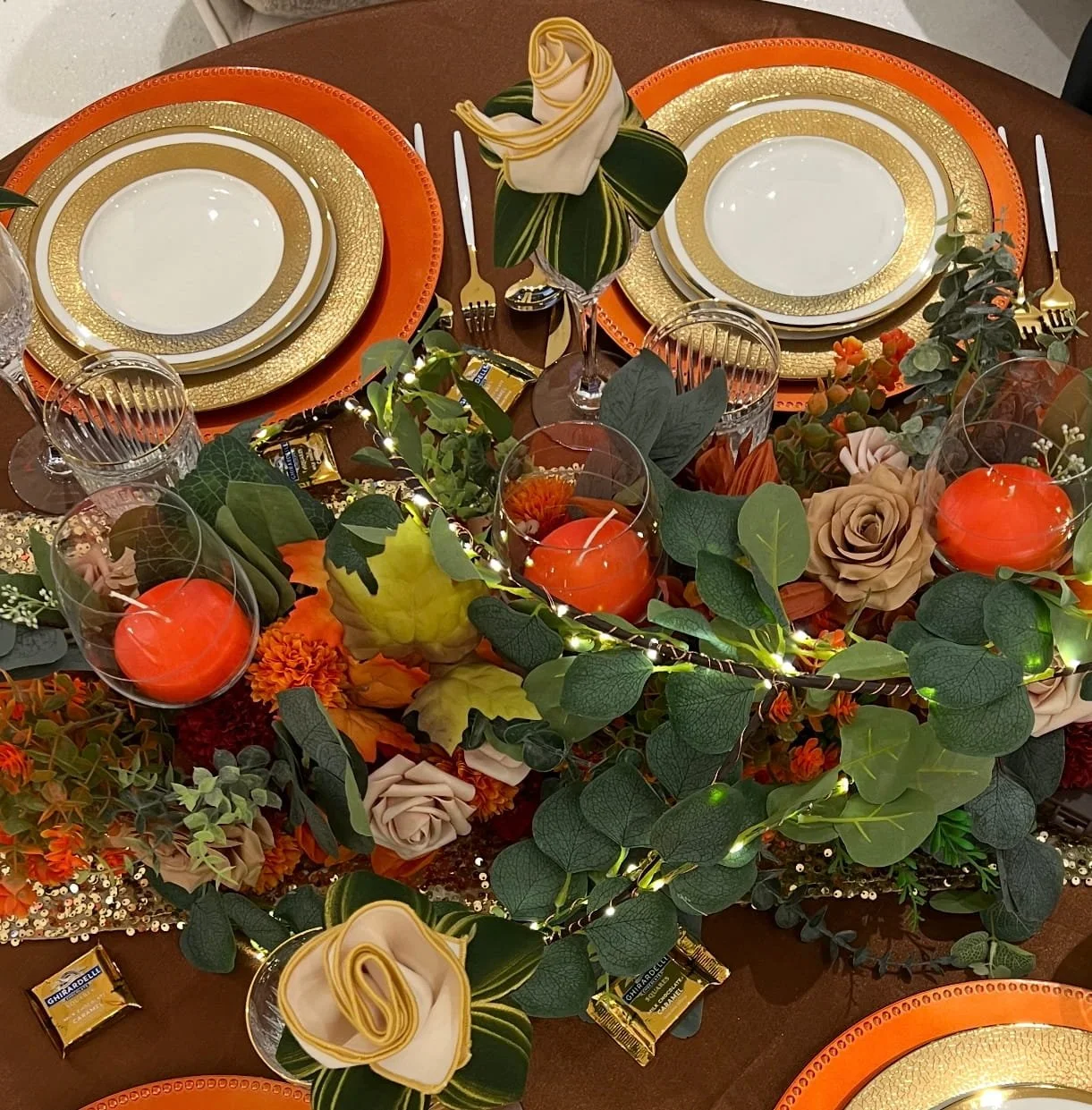 Table setting with orange and gold plates, clear glasses, gold cutlery, decorative candles, and a centerpiece of green foliage, flowers, and string lights.