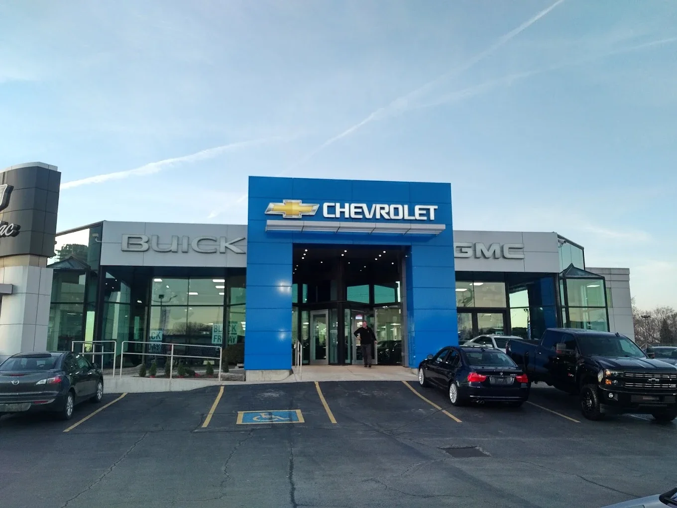 Chevrolet dealership building with Buick and GMC signage, several cars parked outside, clear sky, and a person standing at the entrance.