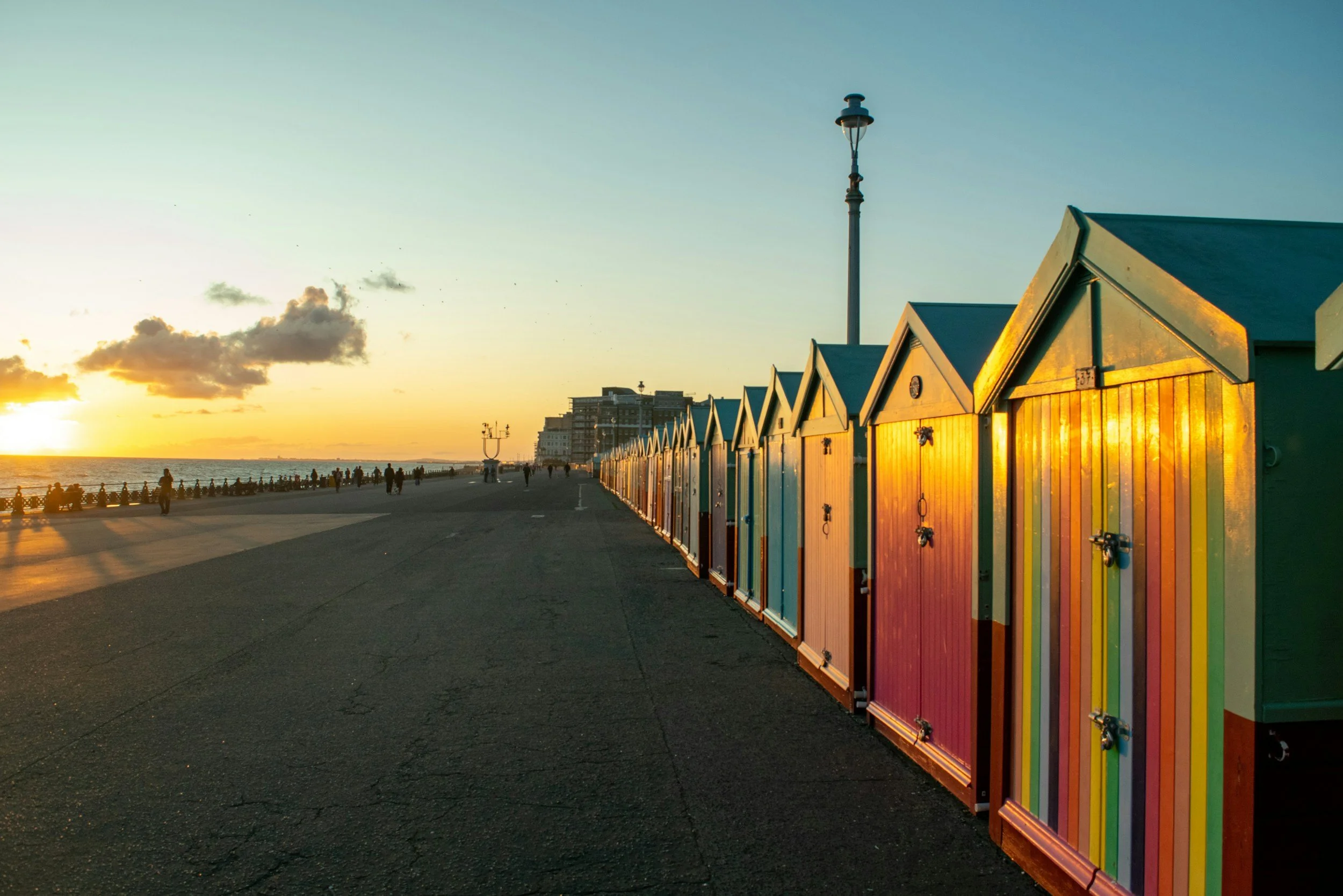 Colorful beach huts lined up along a boardwalk during sunset with a lamppost and buildings in the background.