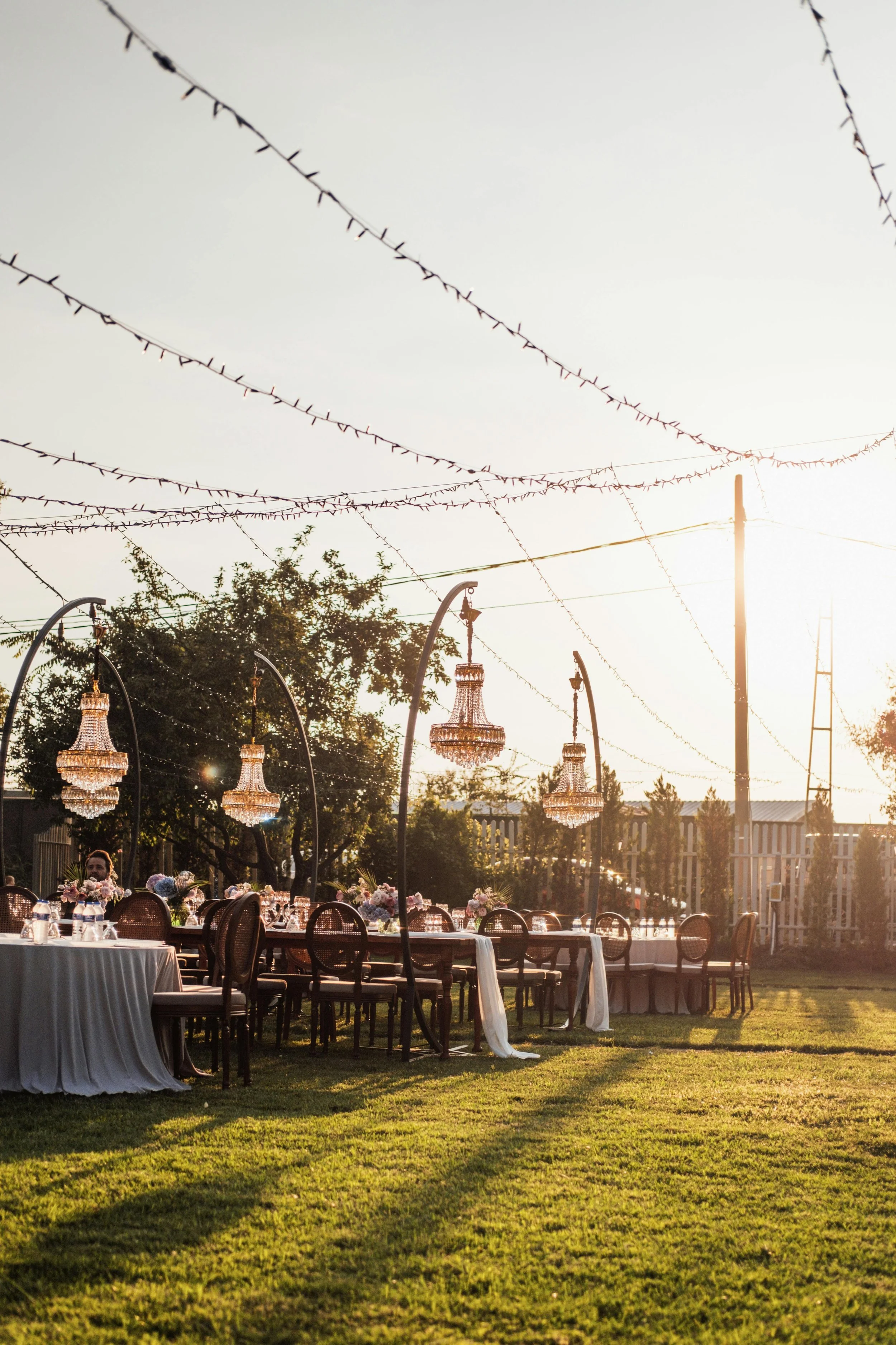 Outdoor event setting with decorated tables, chandeliers hanging from wires, trees, and string lights in the late afternoon or early evening sunlight.