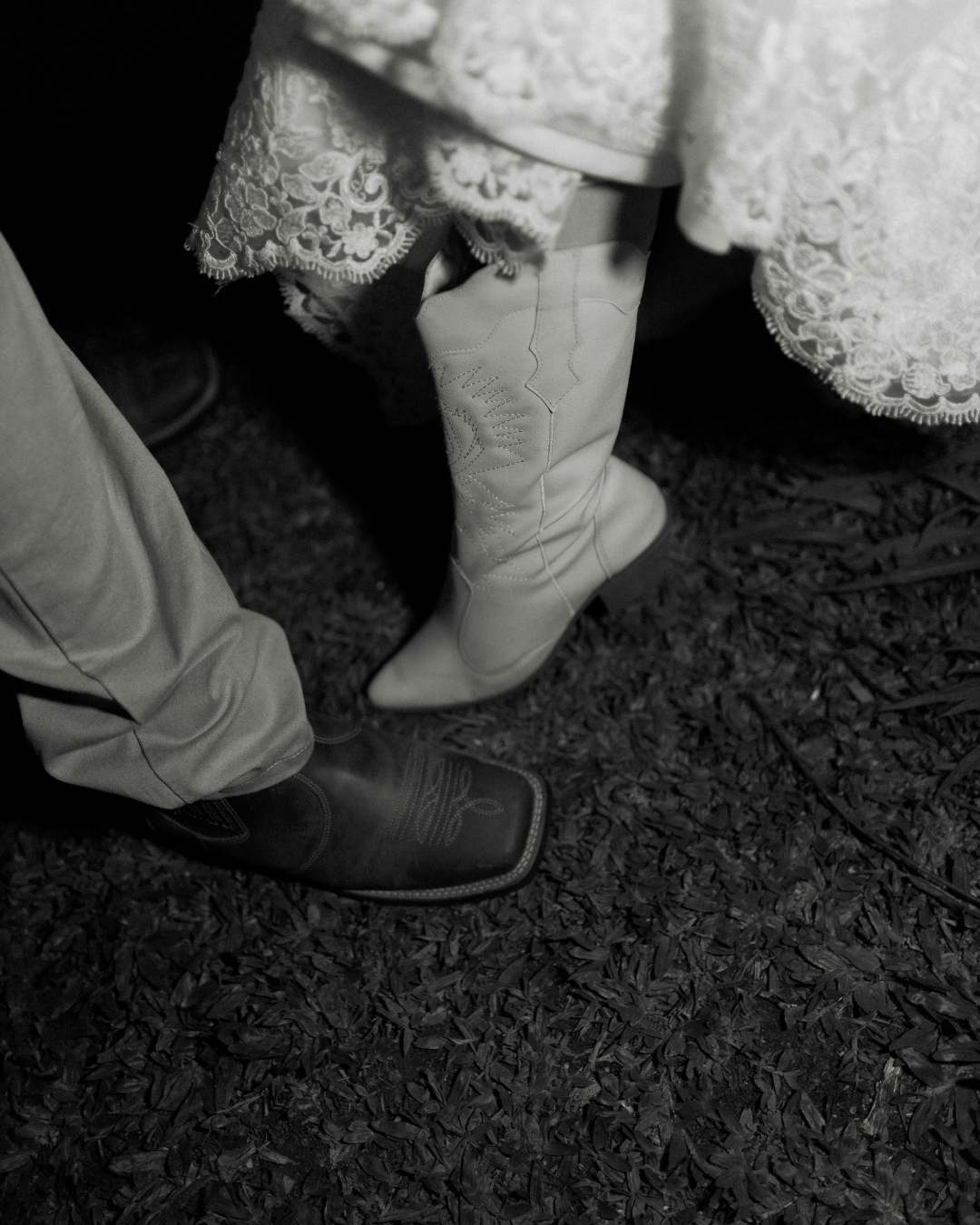 Close-up of two people standing on a ground covered with wood chips, showing their lower legs and feet. One person is wearing light-colored cowboy boots with embroidery, and the other is wearing dark cowboy boots with stitching, with lace and fabric details visible in the background.