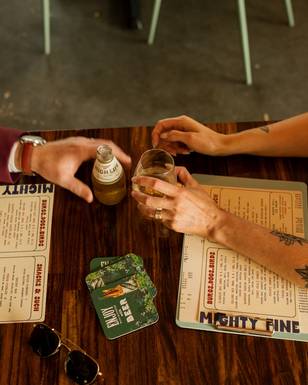 Two people sitting at a wooden table, one holding a beer bottle and the other holding a glass of beer, with menus and sunglasses on the table.