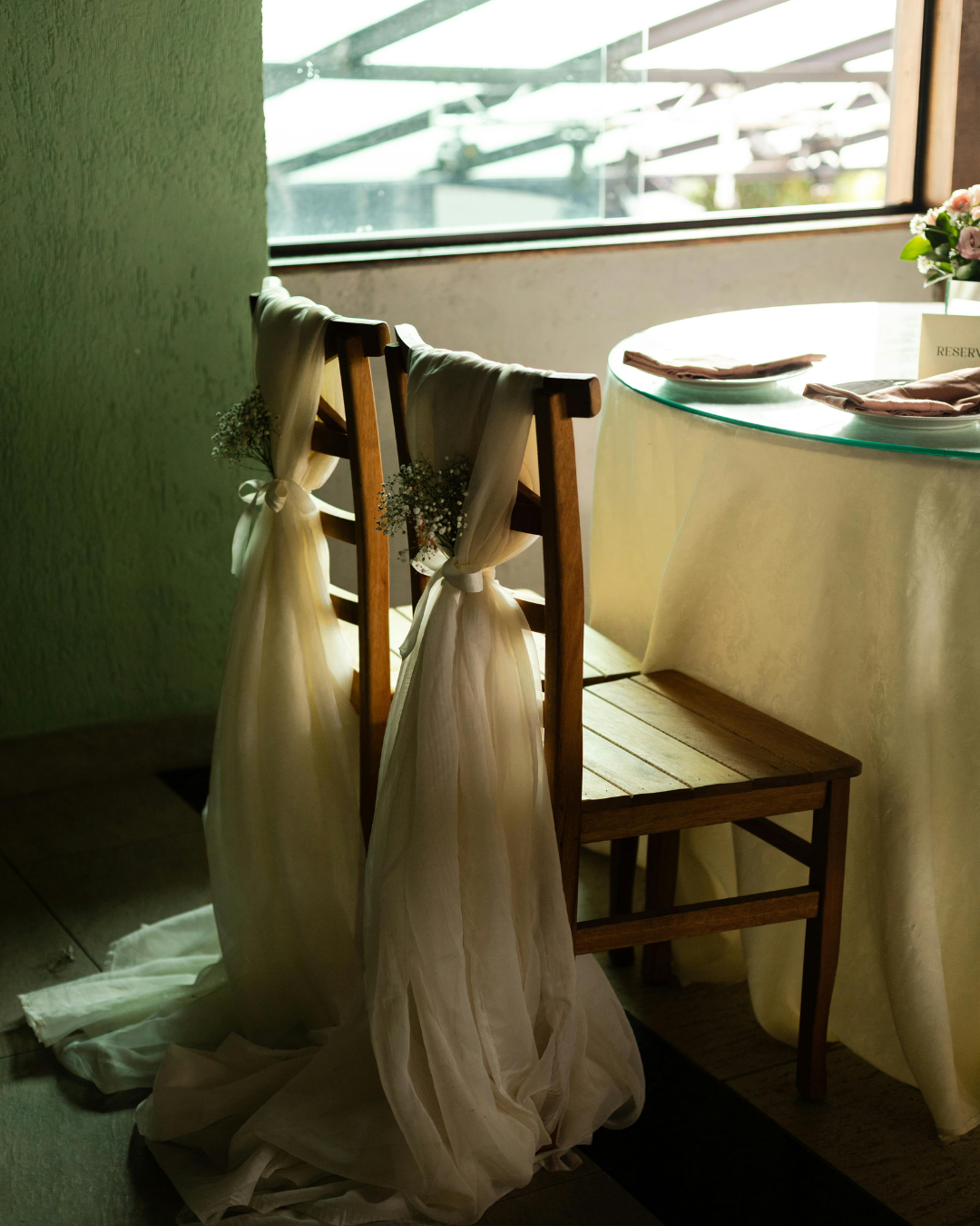Empty wooden chair decorated with white fabric and baby's breath flowers, next to a table with a glass top set with cloth napkins in a softly lit room.