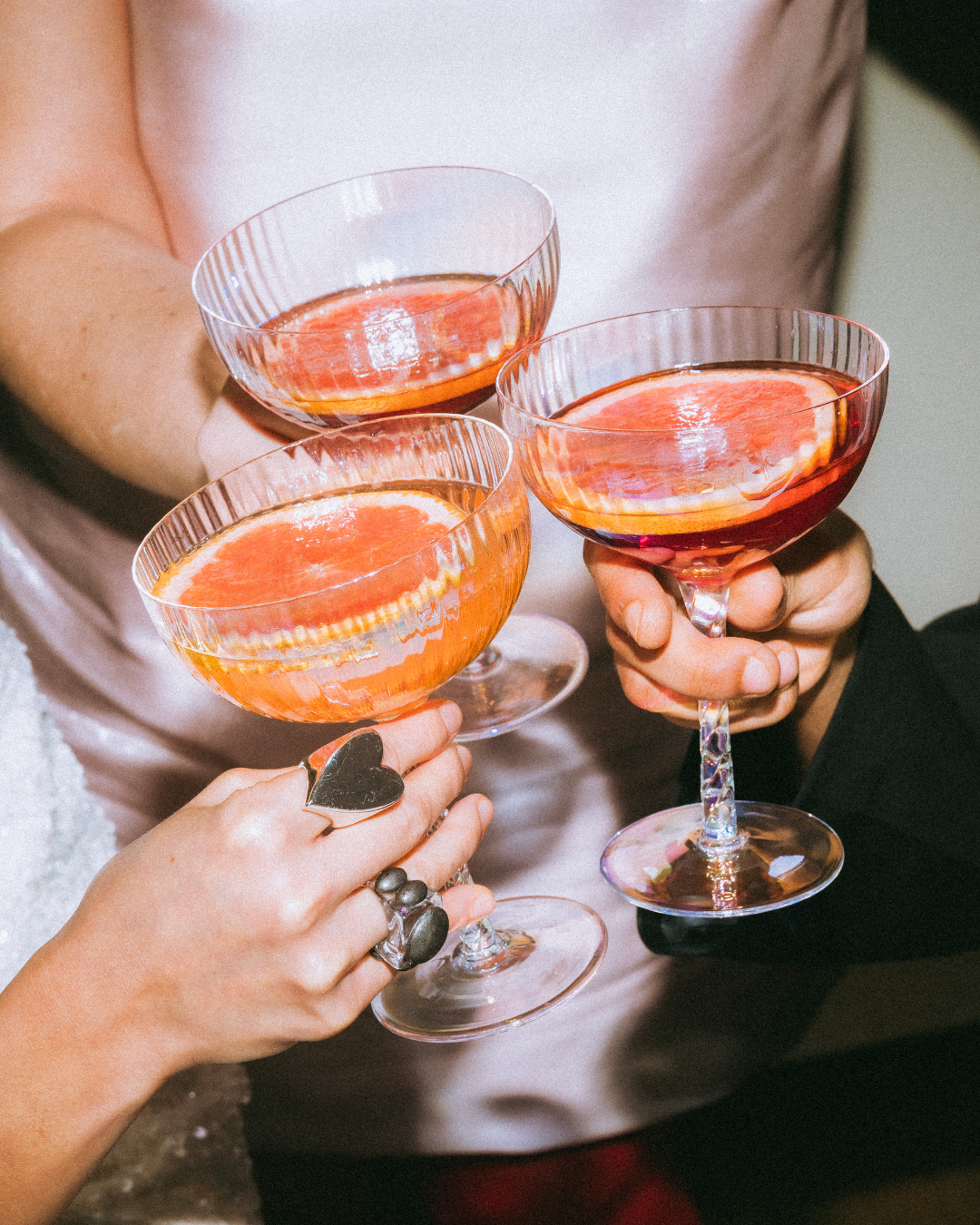 Three people holding wine glasses filled with a pink-colored drink and slices of grapefruit inside.