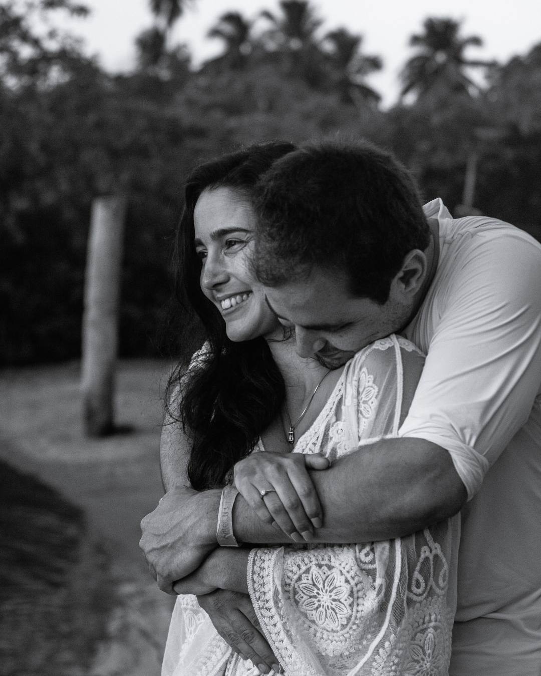 A black and white photo of a man hugging a woman from behind, both smiling, outdoors with trees in the background.