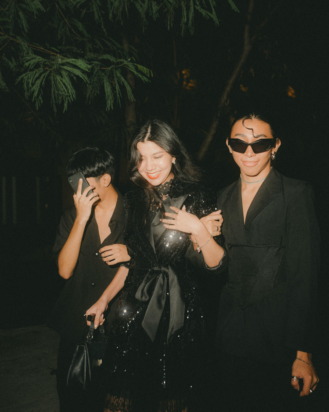 Three stylish women dressed in black at night, one with sunglasses, one smiling with hand on chest, one talking on phone, standing outdoors with trees in background.