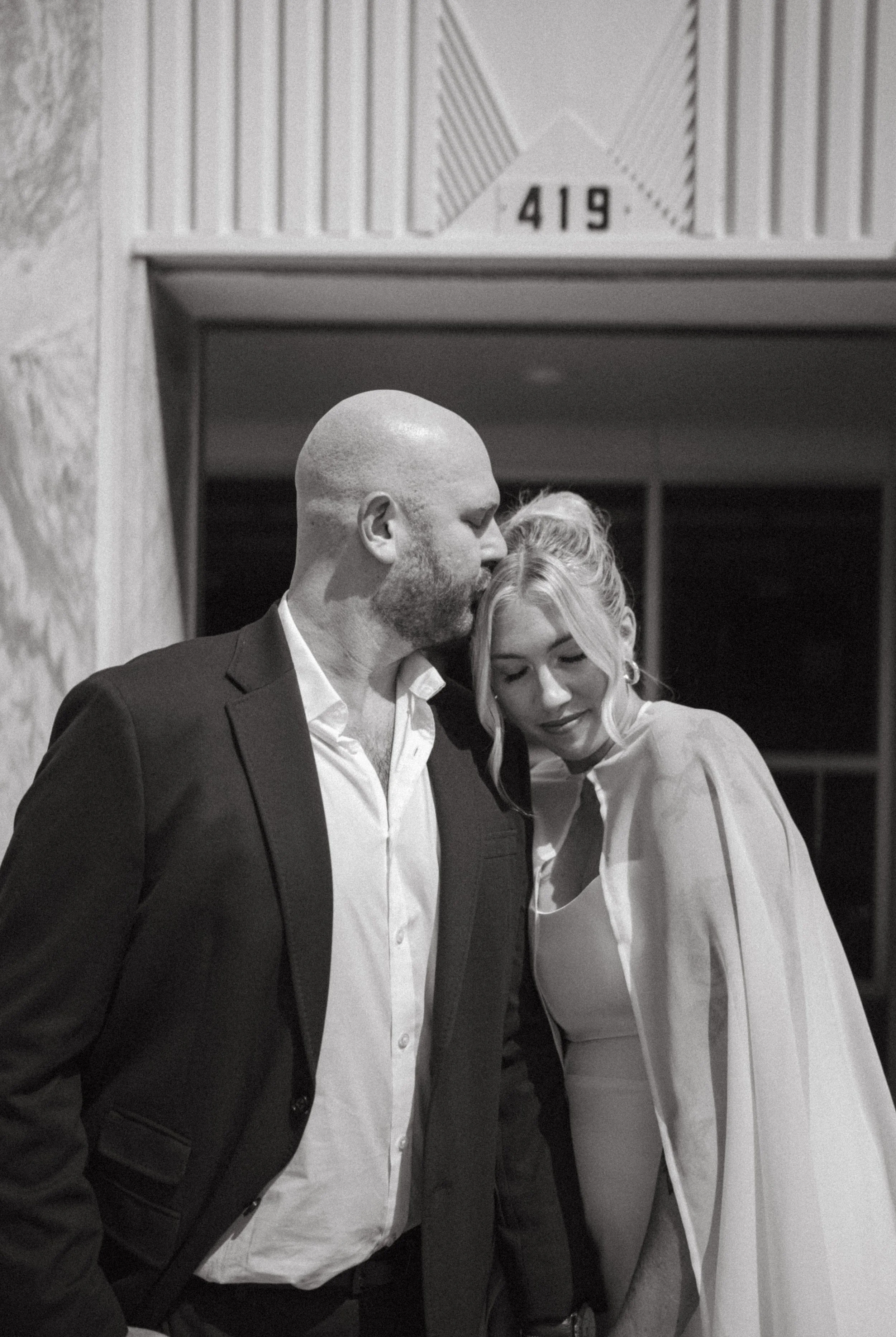 A man in a tuxedo and a woman in a formal dress are leaning their heads together in an affectionate pose.