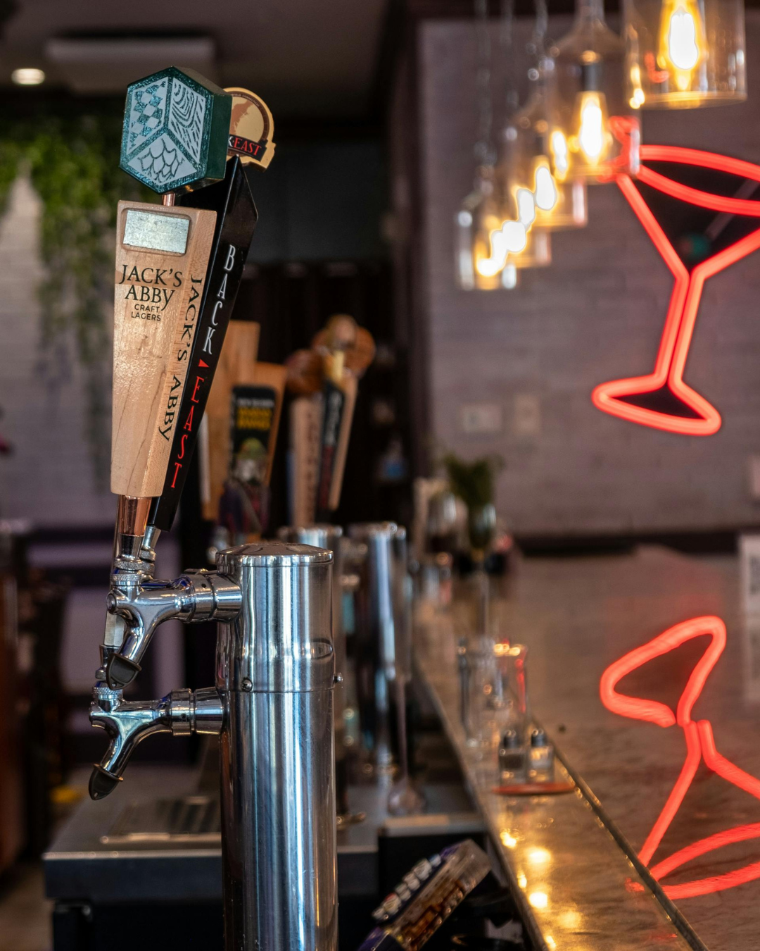 Close-up of a bar with beer taps, including Jack's Abby craft lagers, against a blurred background with neon lighting and bar decor.