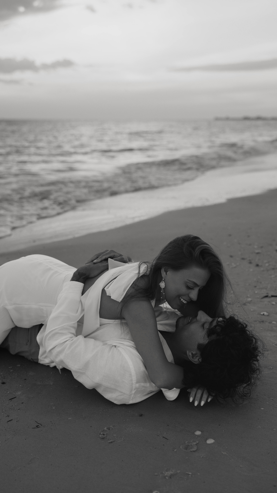 A black and white photo of a smiling woman on top of a man lying on a beach, with the ocean in the background.