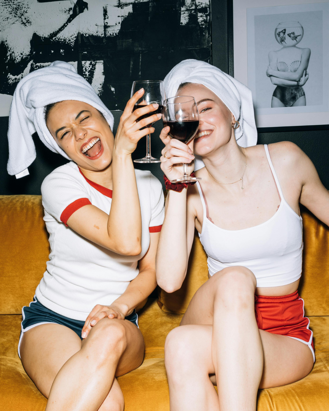 Two women with towels on their heads sitting on a yellow couch, enjoying glasses of red wine, smiling and having fun.