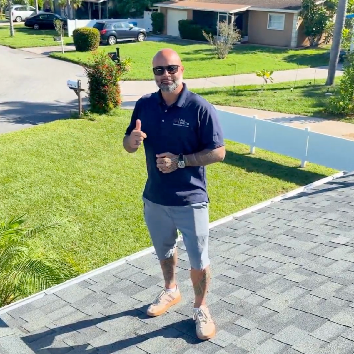 A man standing on a residential driveway, giving a thumbs-up gesture. He is wearing sunglasses, a dark blue polo shirt, light gray shorts, and beige sneakers. The background shows a lawn, a sidewalk, houses, and parked cars on a sunny day.