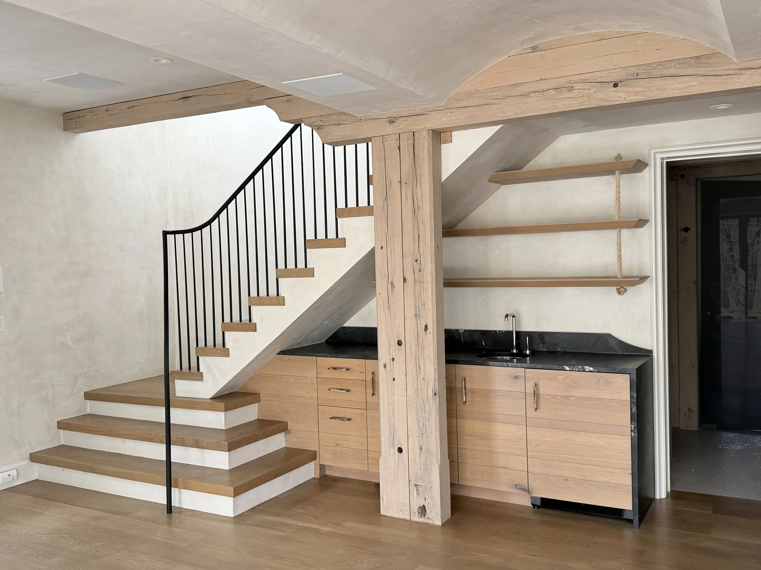 Modern interior of a house with a wooden staircase, black metal railing, wooden shelves on the wall, and kitchen with wooden cabinets and black countertops.
