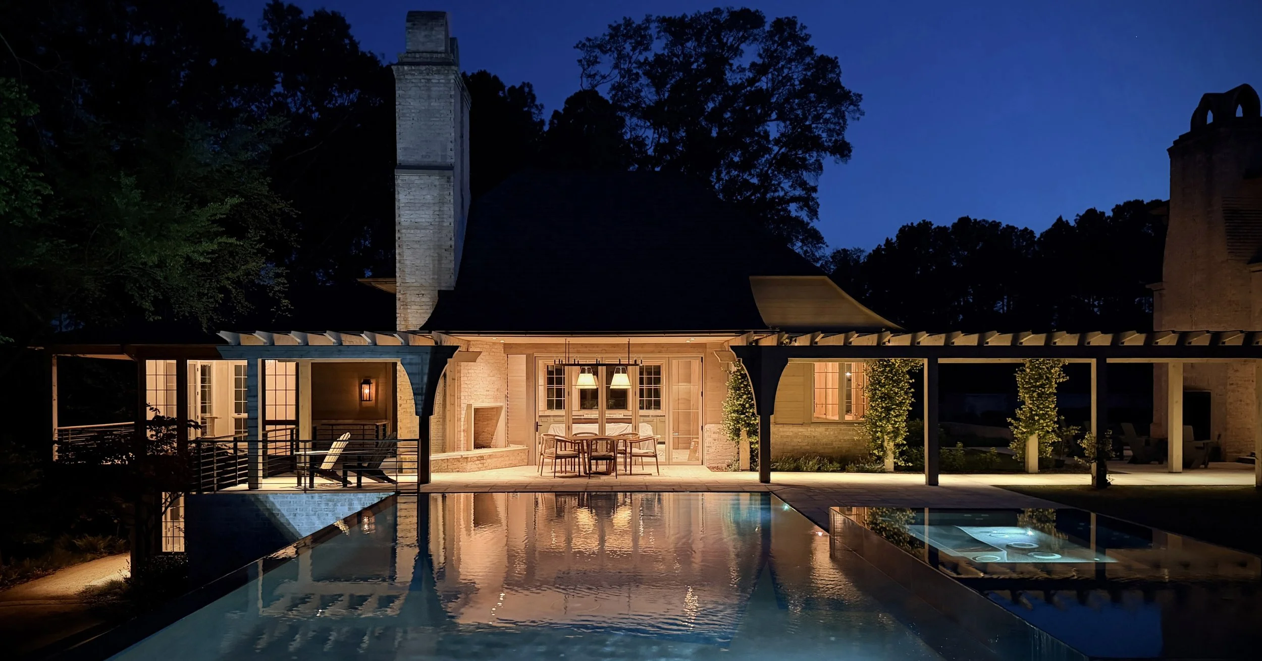 Night view of a house with lit interior, a covered patio, and a swimming pool in the foreground, surrounded by trees.