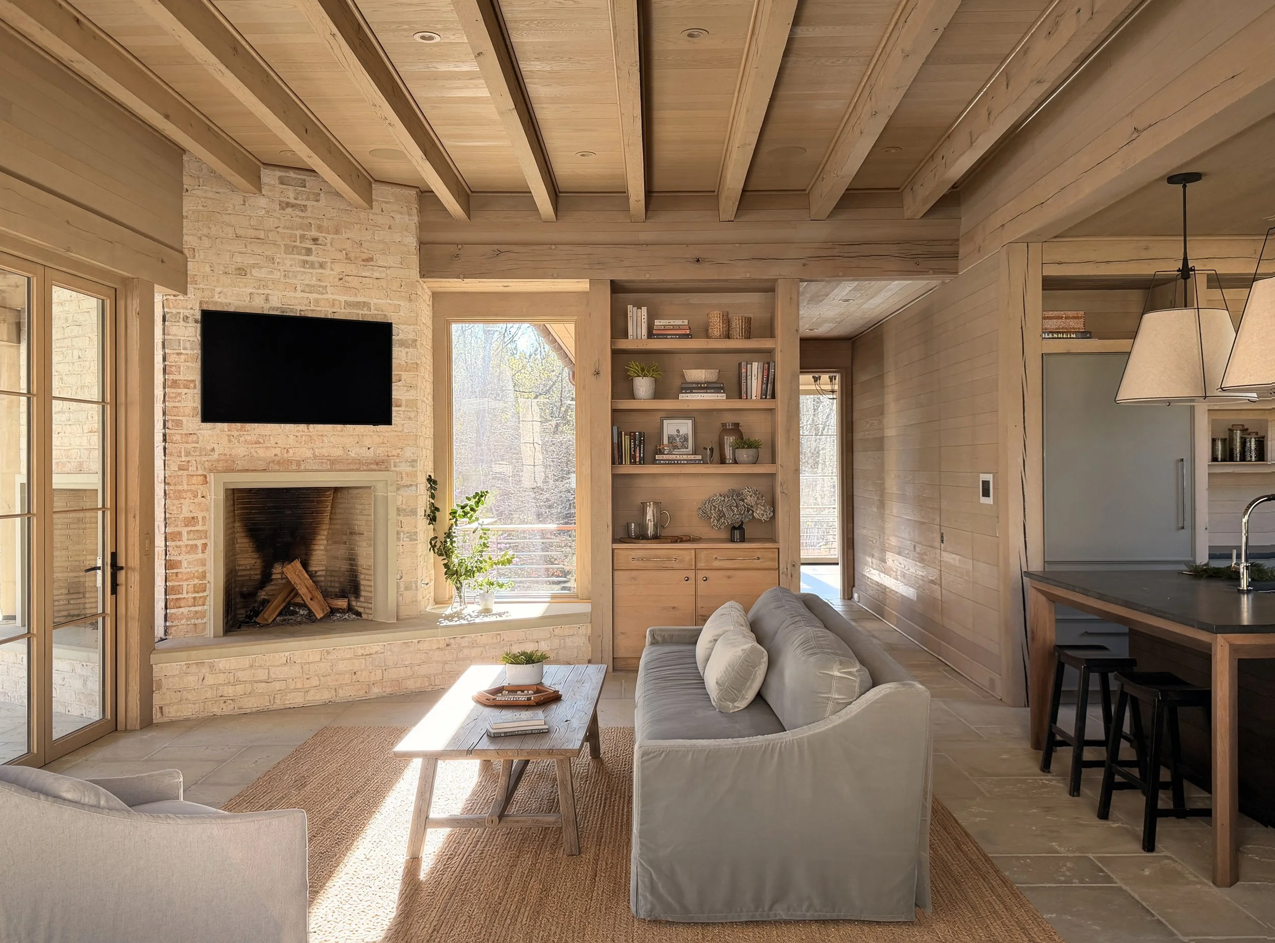 A cozy living room featuring a brick fireplace, a large flat-screen TV, a white sofa with pillows, a wooden coffee table, and built-in wooden shelves with books and decorations. Large windows let in natural light, and a door with glass panes leads ou