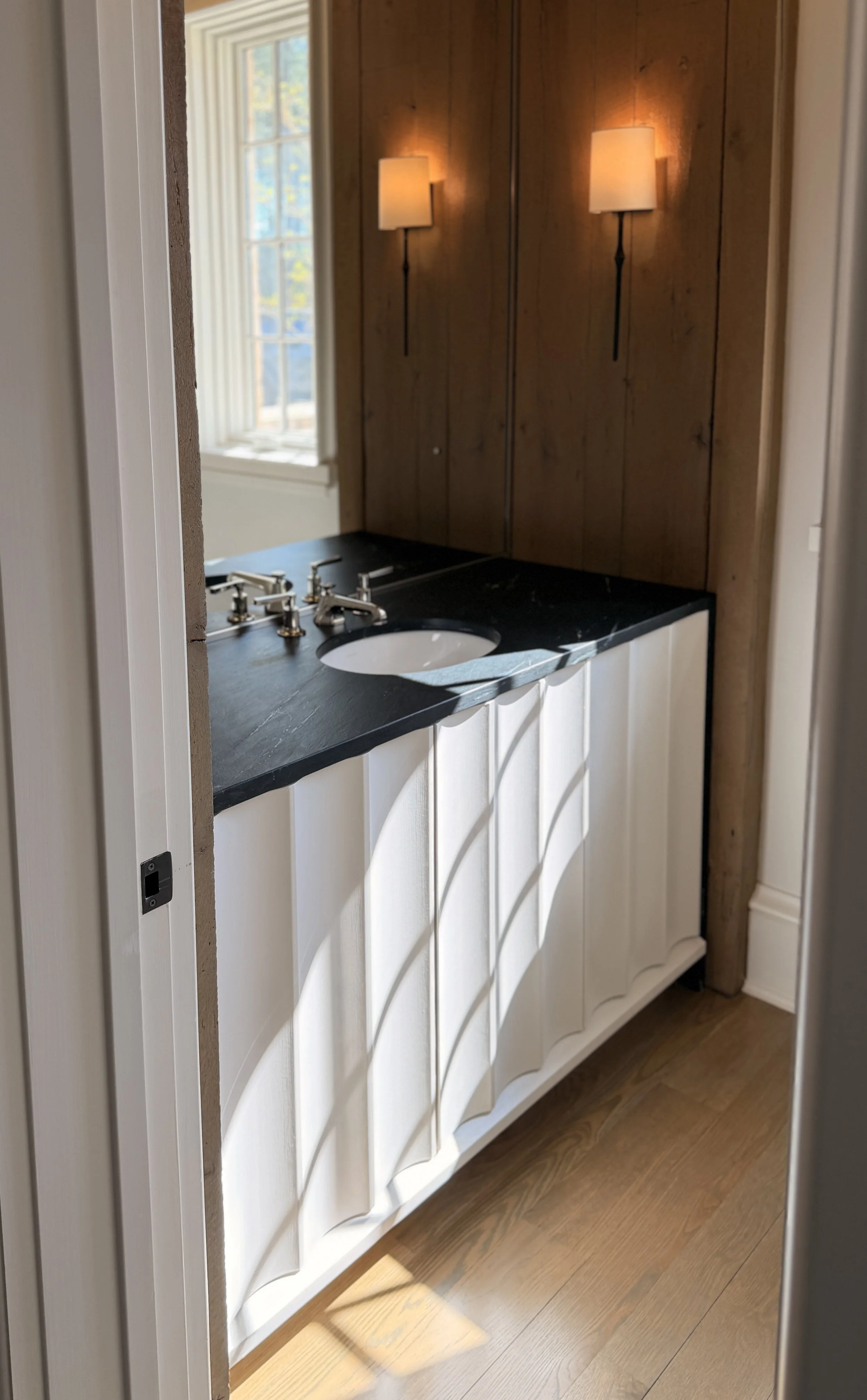 A bathroom vanity with a black countertop, an undermount sink, silver faucets, white cabinet doors with scalloped detailing, wooden flooring, a window with vertical blinds, and wooden wall panels with two wall-mounted lamps.