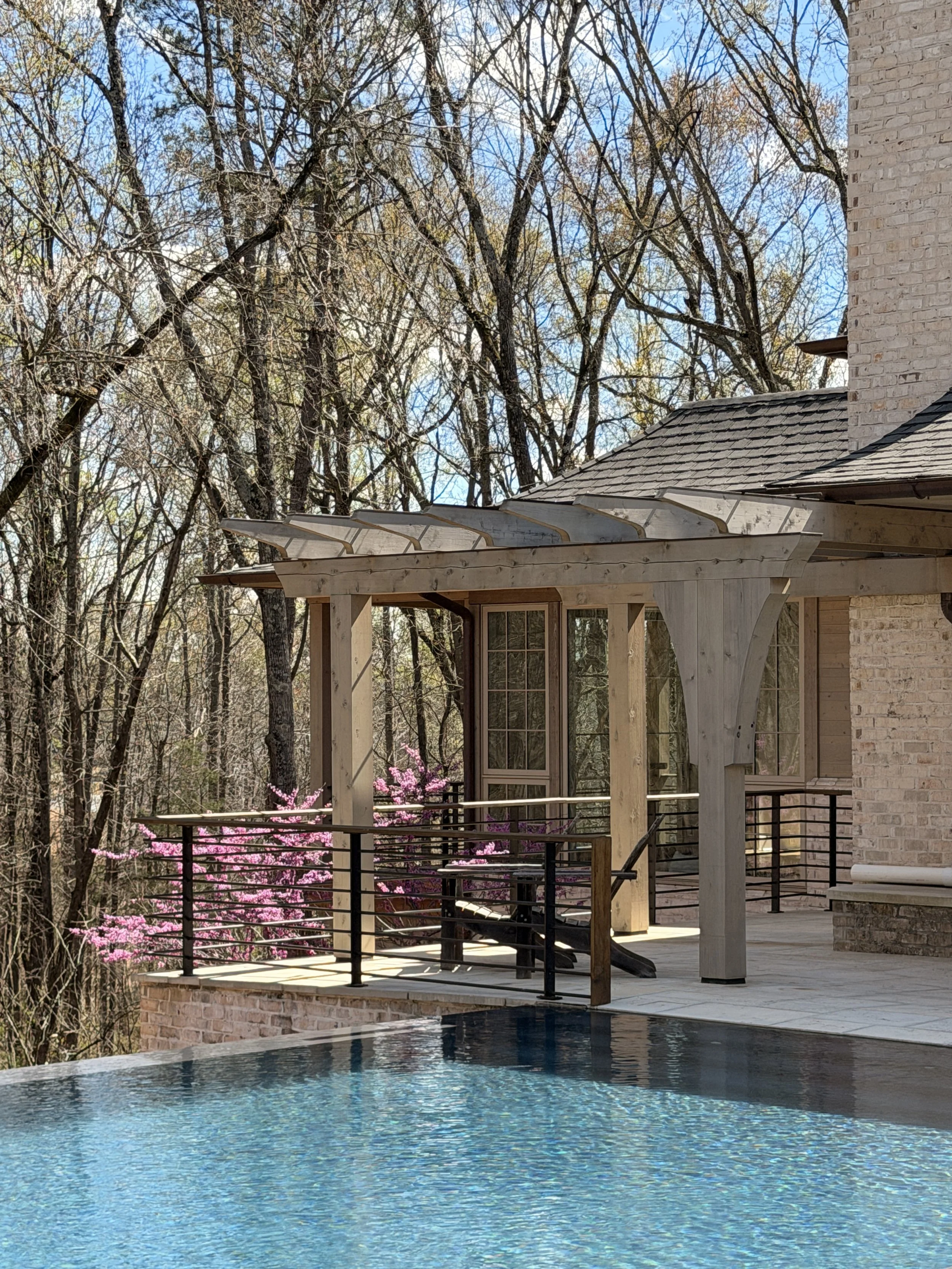 A backyard patio with a wooden pergola, overlooking a swimming pool with blue water. There are pink flowering bushes and tall trees with bare branches, indicating early spring or late fall. Part of a brick house is visible on the right.