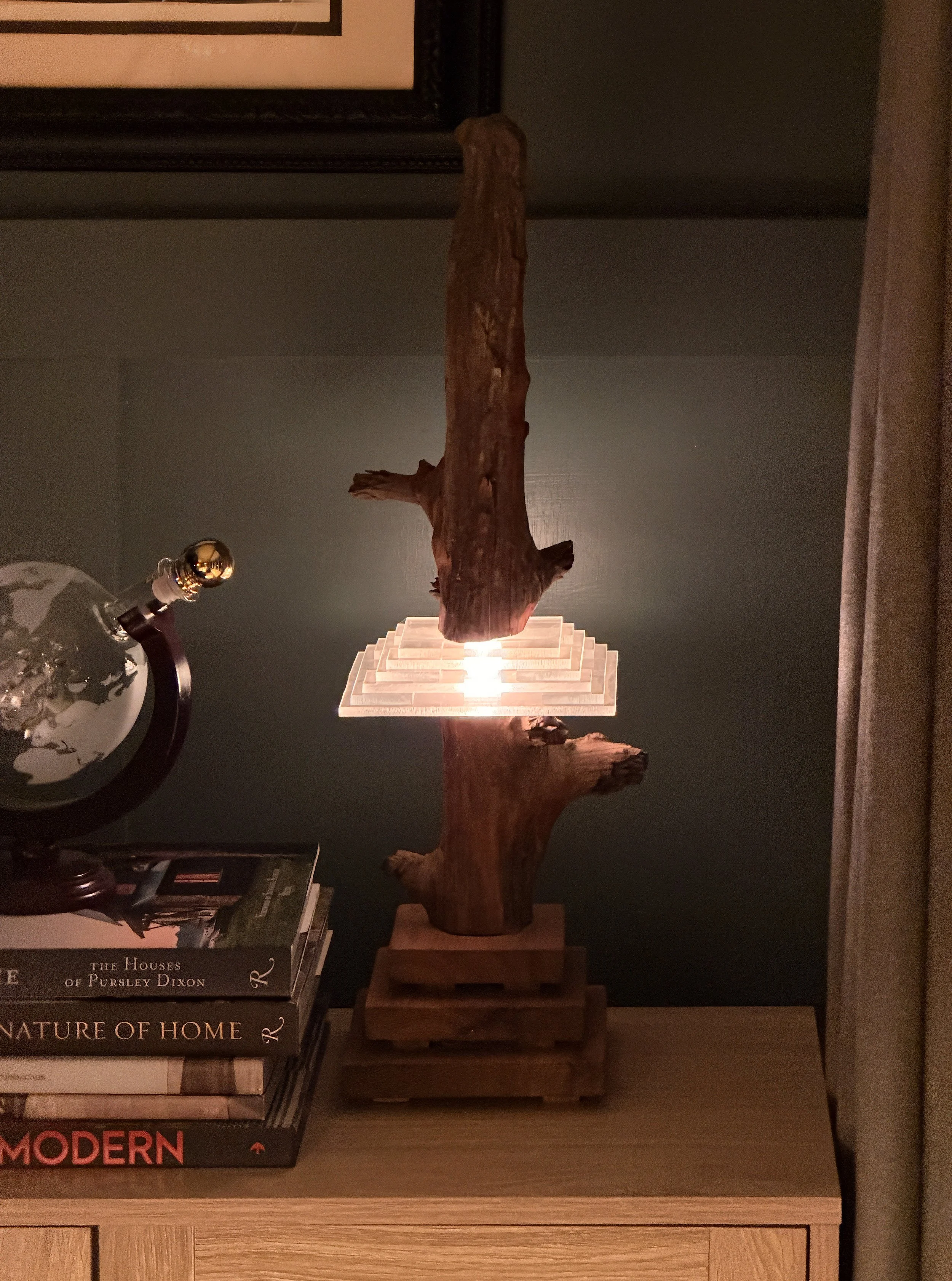A wooden sculpture resembling a tree trunk with branches, placed on a lit stepped platform against a dark wall. To the left, a globe lamp and some stacked books are visible.