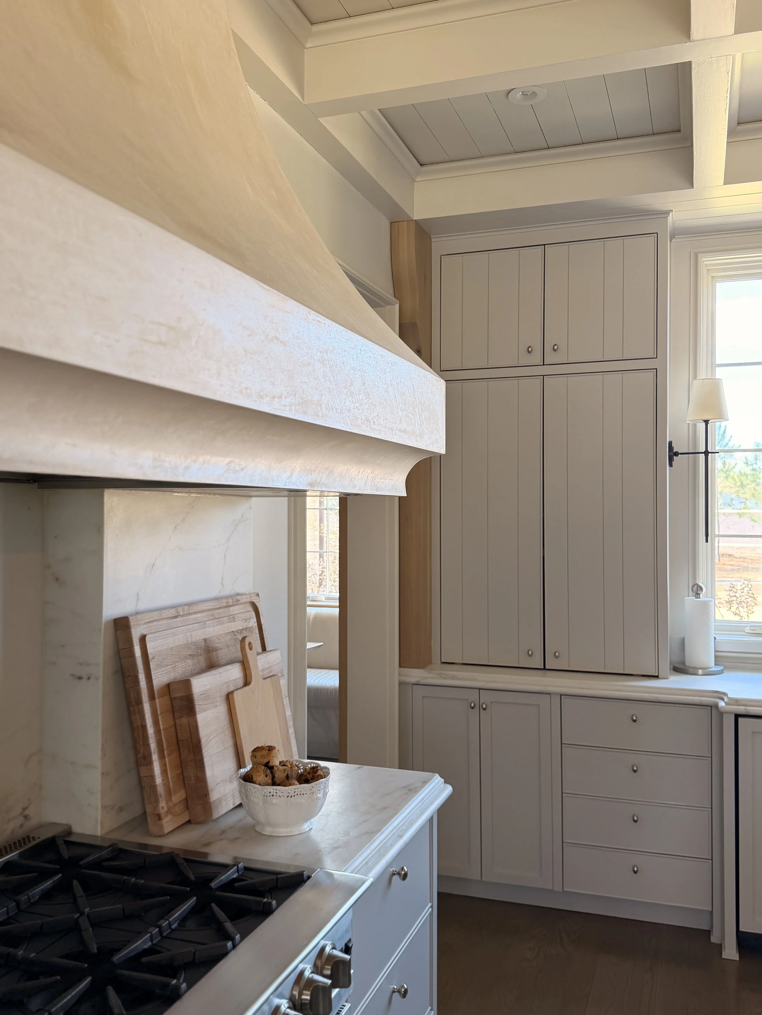 A kitchen with white cabinetry, a marble countertop, and a small table with a bowl of cookies and wooden cutting boards. A large window lets in natural light, with a lamp and paper towel roll nearby.