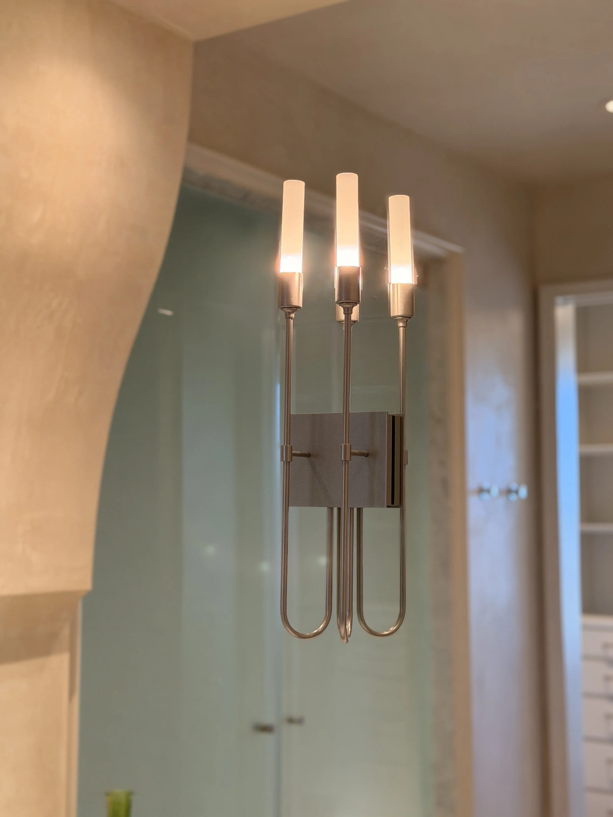 Three silver-colored metal hooks hanging from a wooden surface, with a blurred background of a room interior with beige walls and a white door.
