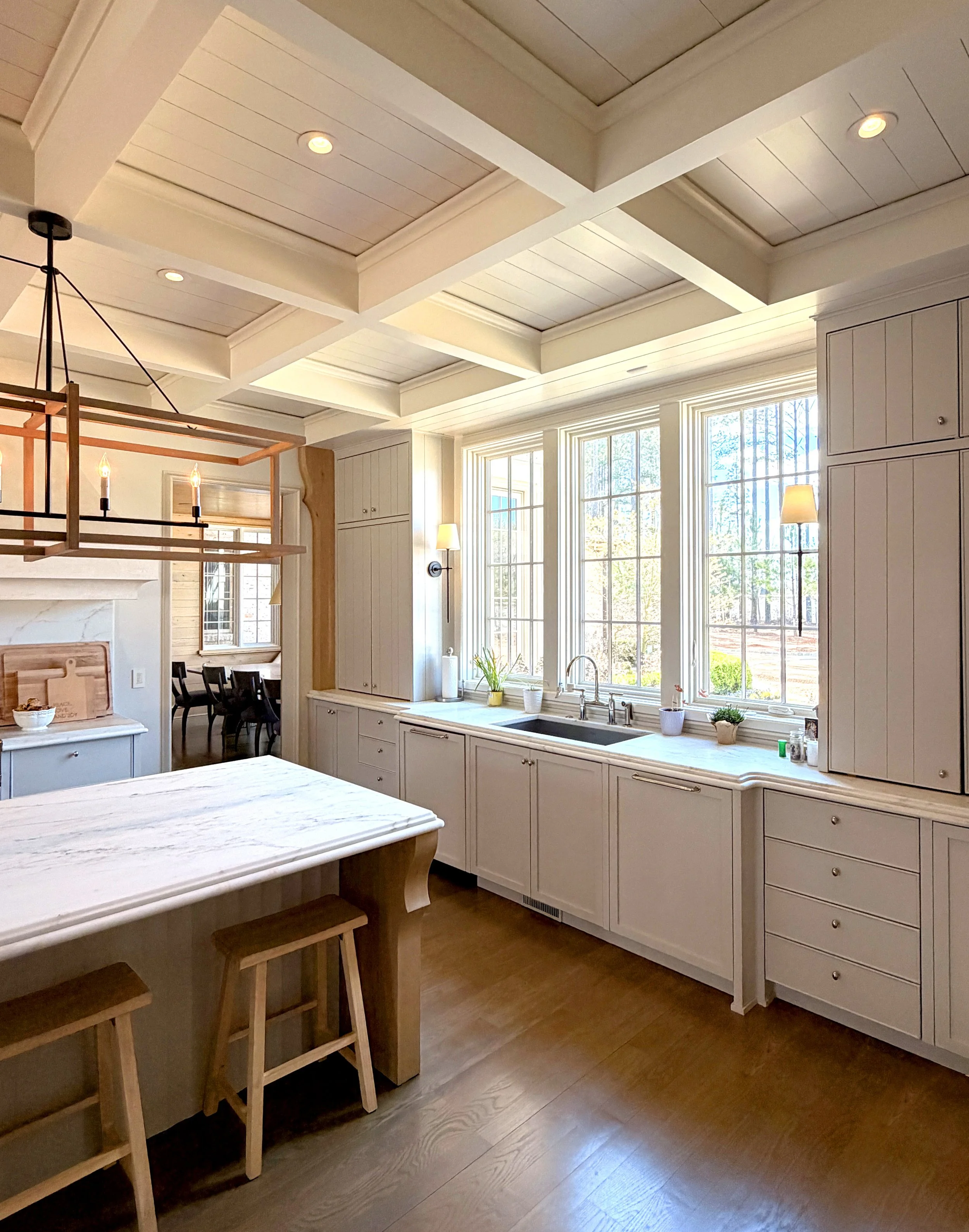 A bright kitchen with large windows and white cabinetry, featuring a marble countertop, potted plants, and a dining area with black chairs. A wooden bar with stools and a modern chandelier are visible.