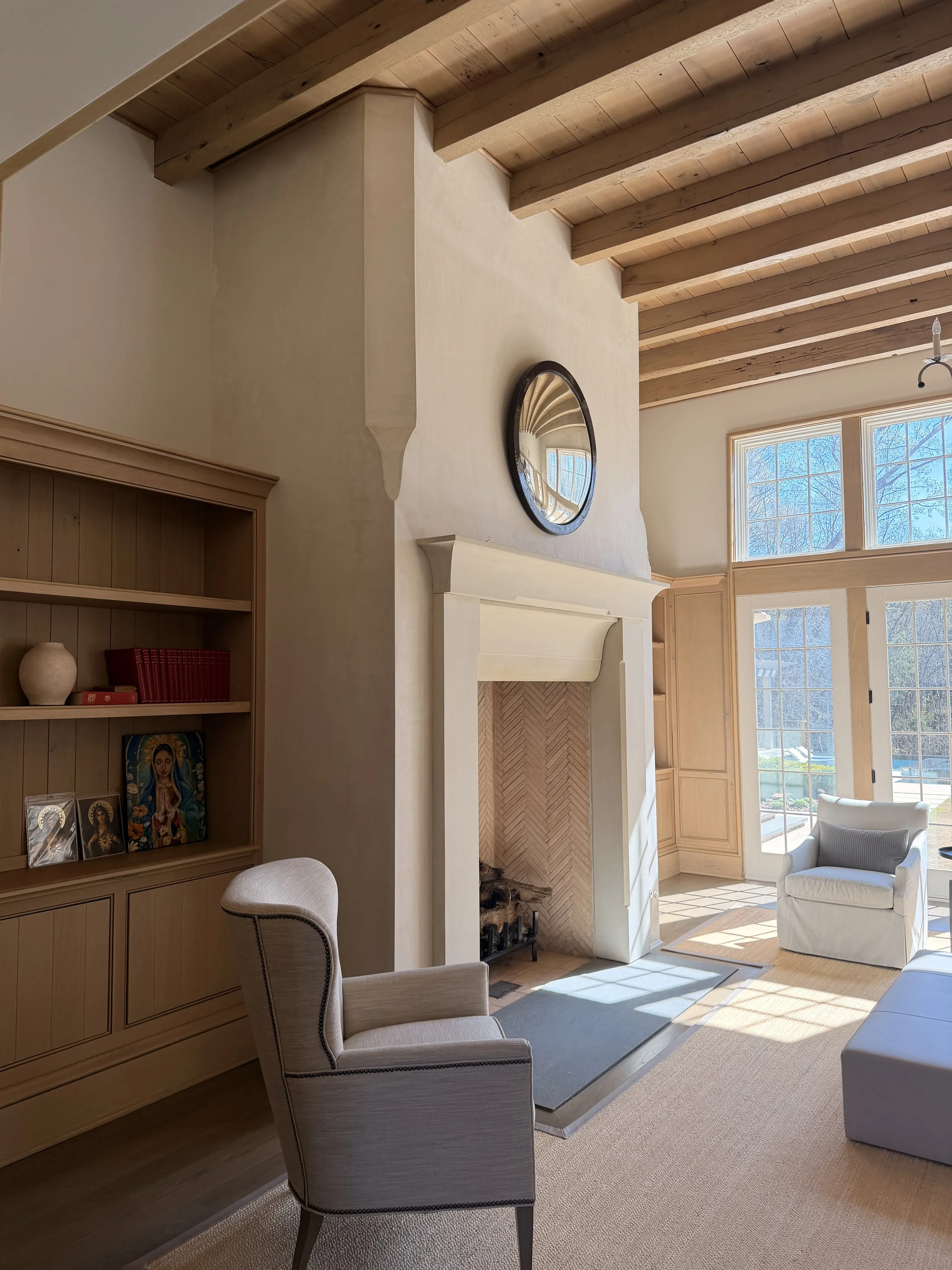 A cozy living room with a stone fireplace, a large window letting in natural light, an armchair, a bookshelf with books and decor, and a ceiling with wooden beams.