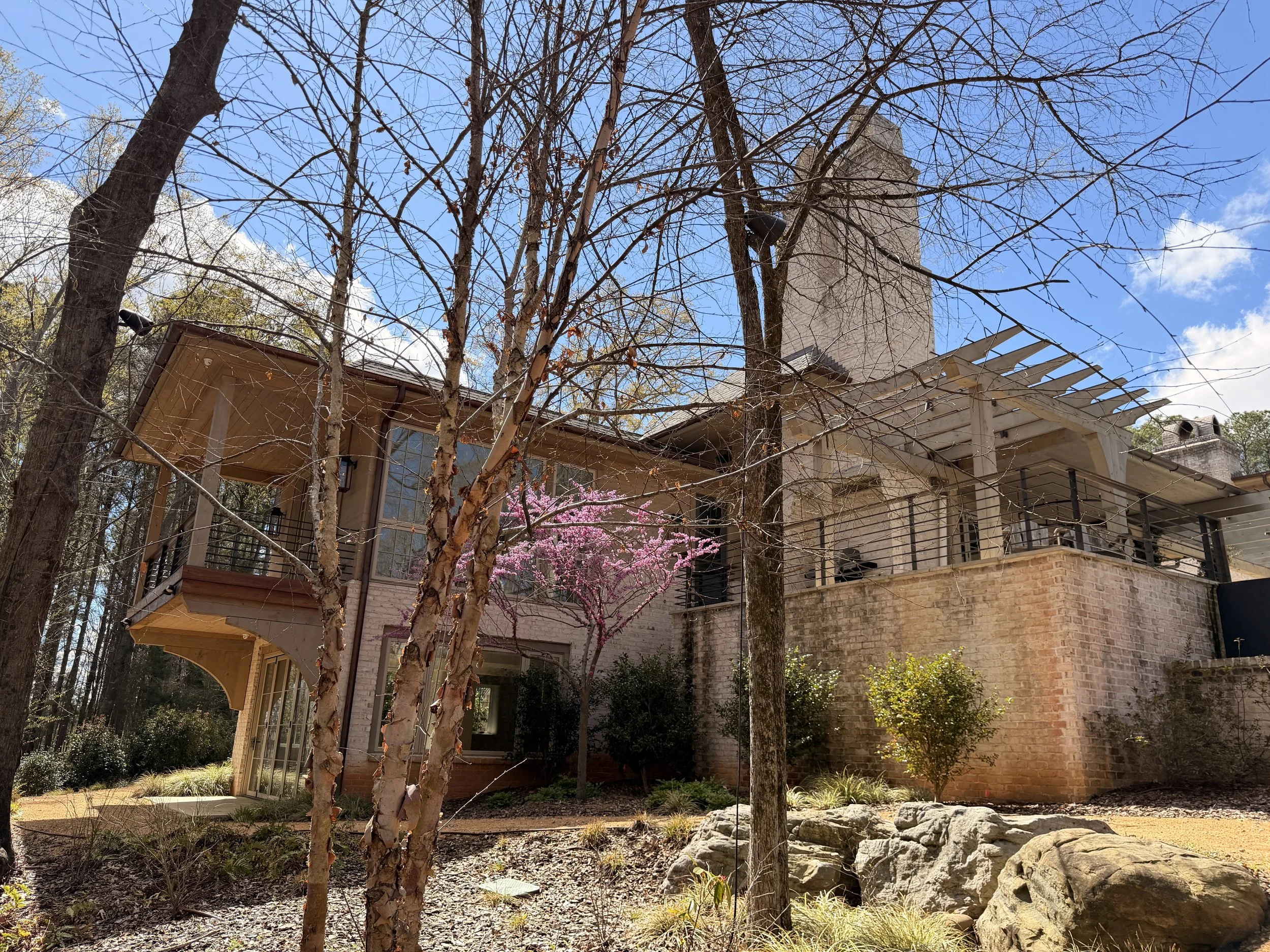 View of a house with a brick exterior, multiple balconies, and a chimney in a wooded area with leafless trees, pink flowering tree, and rocks in the yard under a blue sky.