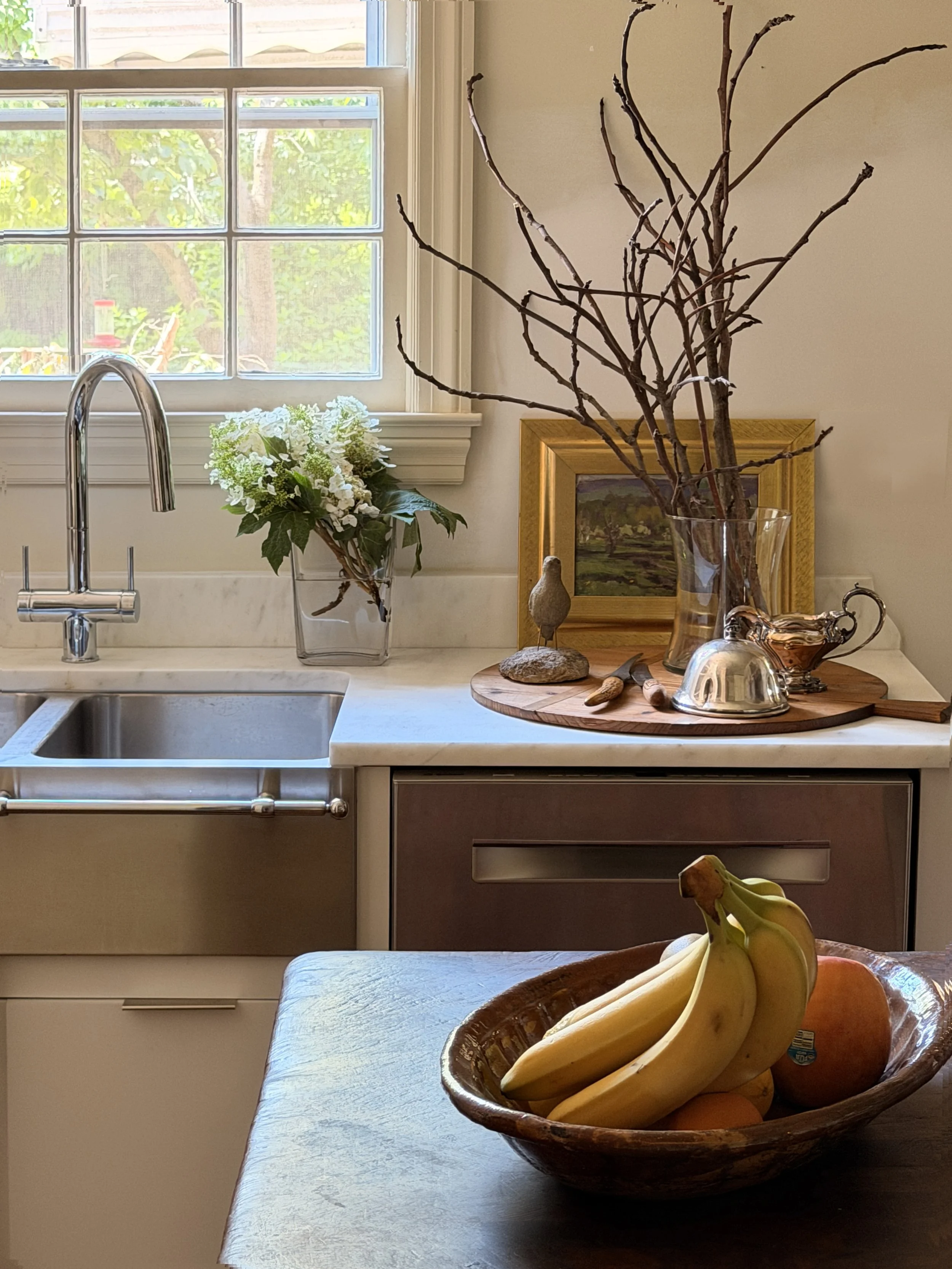 A kitchen countertop with a bowl of bananas, an apple, and a orange. In the background, a window, a vase with white flowers, and decorative branches in a glass vase. A framed landscape painting and a small bird figurine are also on the counter.