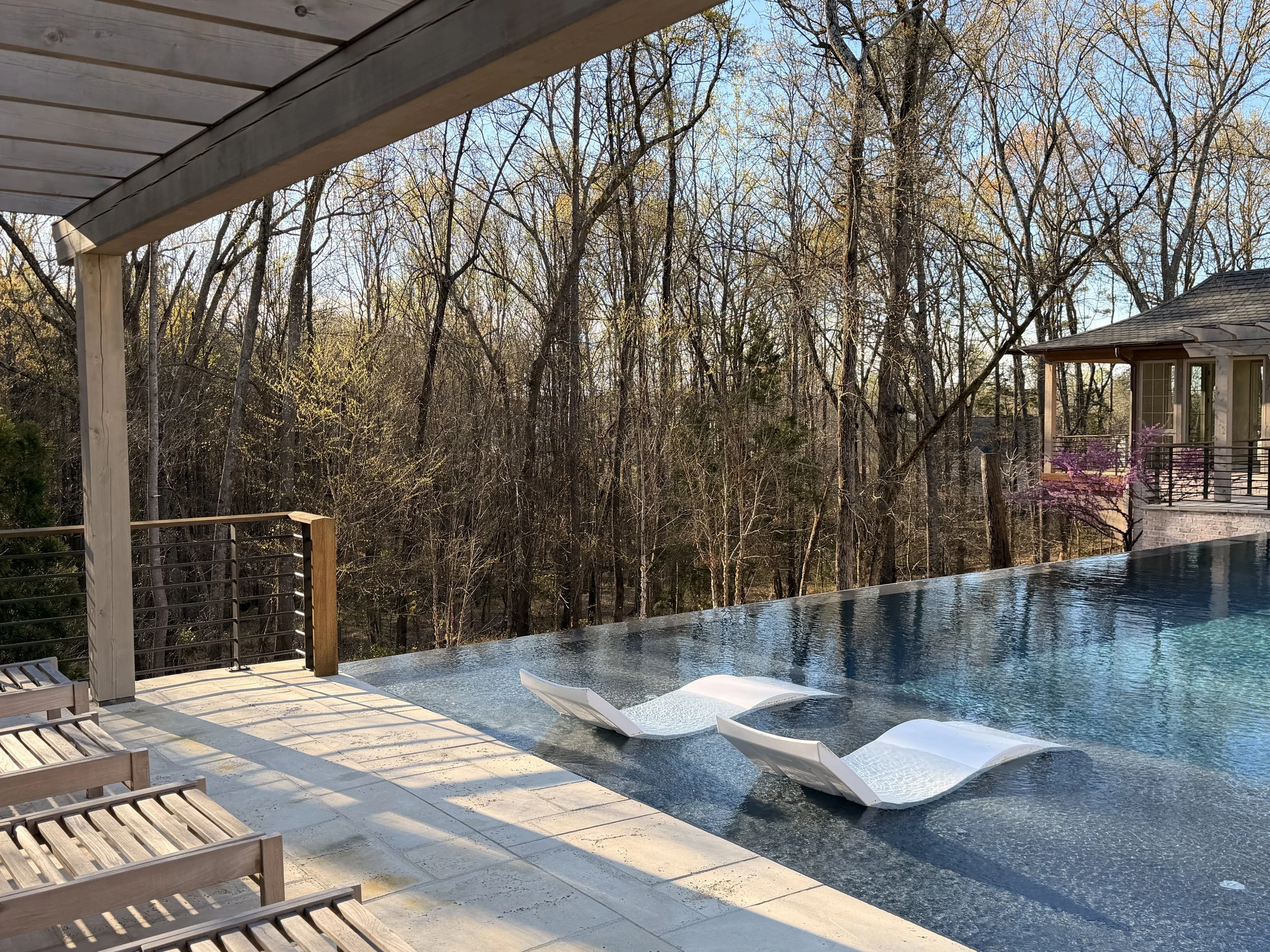 View of a backyard with a swimming pool, surrounded by a wooden deck with lounge chairs, and a view of tall trees with bare branches and a neighboring house with a porch, in the late afternoon sunlight.