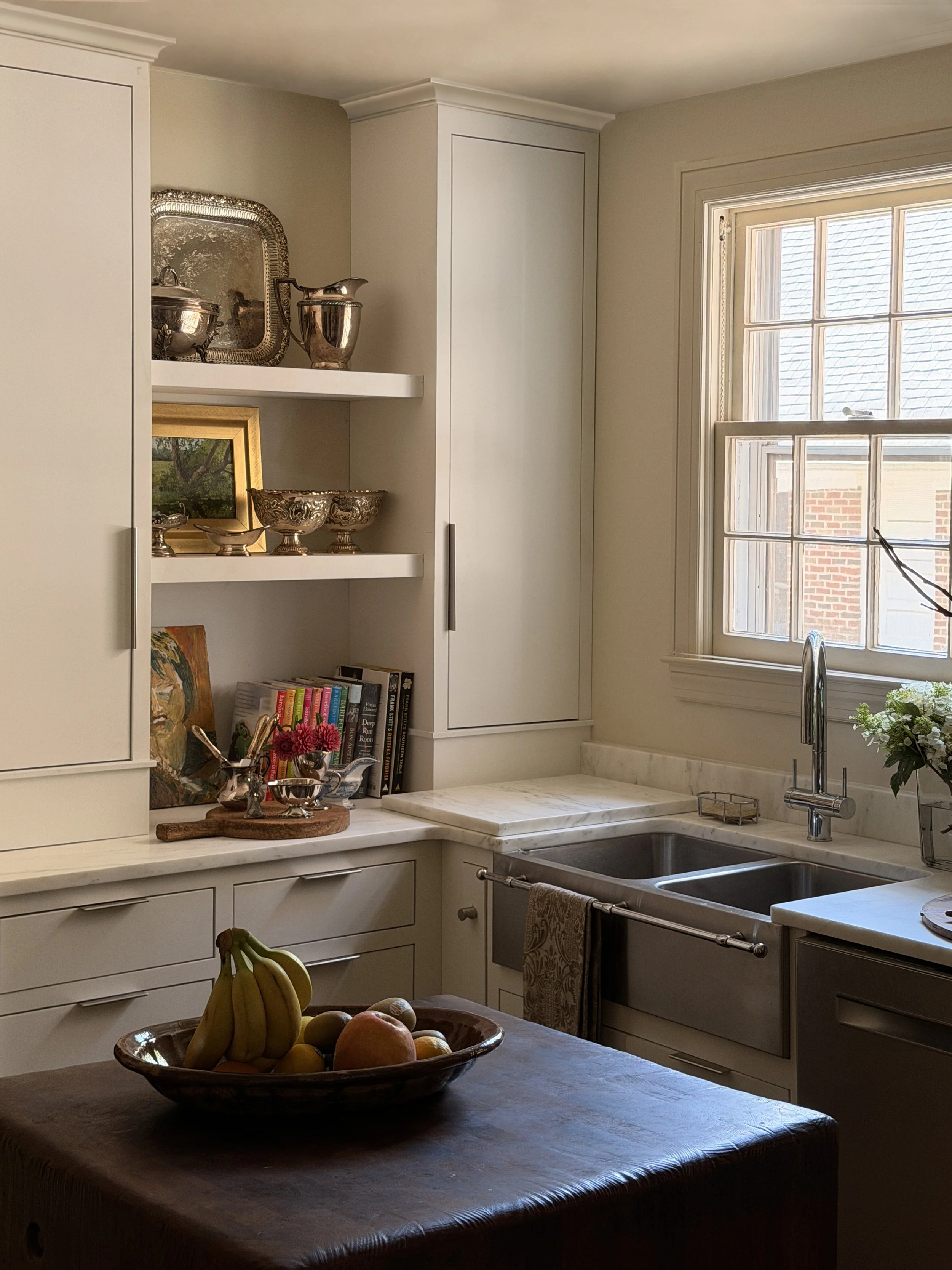A kitchen with white cabinets, a window above the sink, decorated with silverware, framed art, and books on the counter, and a wooden fruit bowl on the table.
