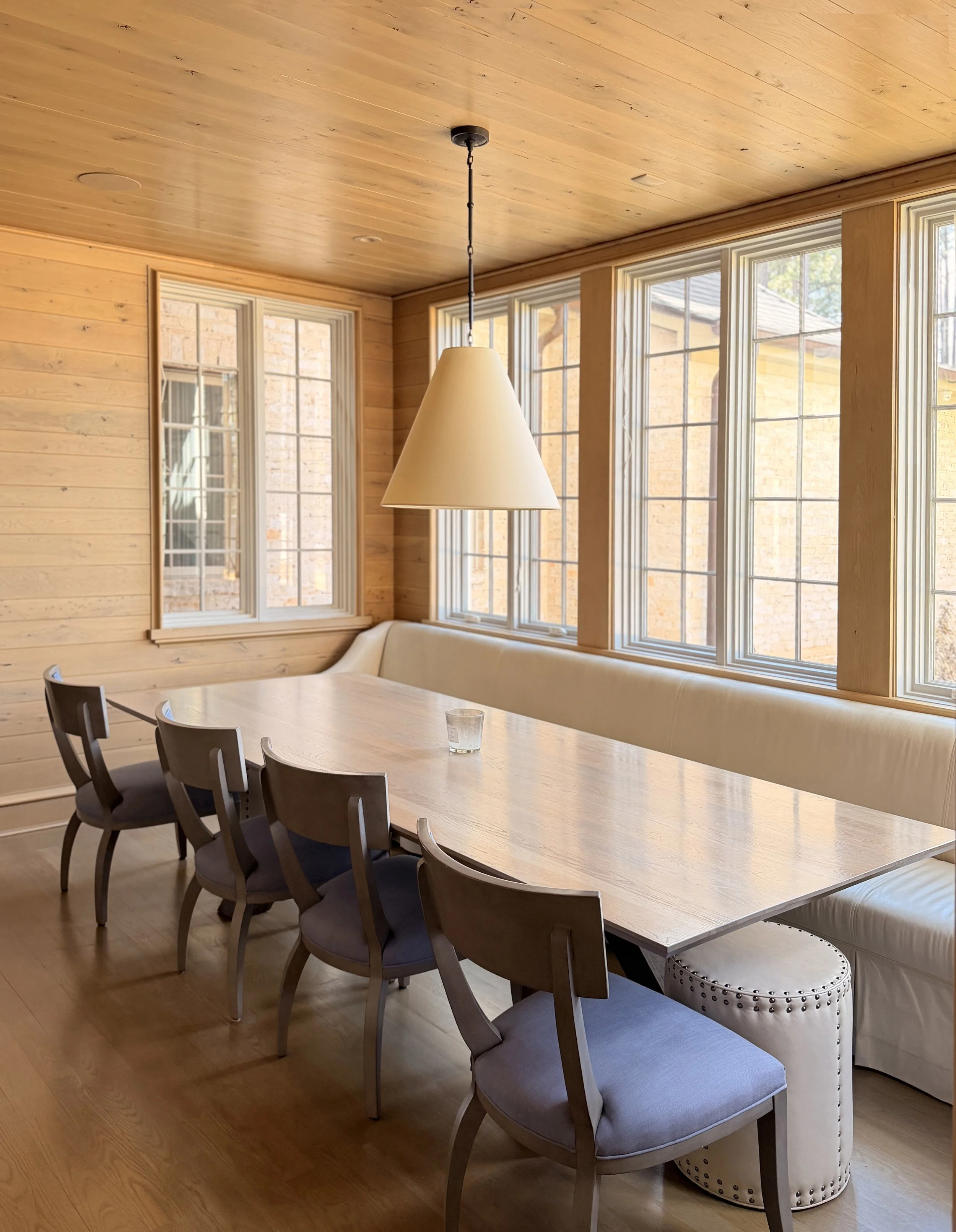 A dining room with a light-colored wooden ceiling and walls, large windows, a long wooden dining table, black chairs with cushioned seats, and a white upholstered side bench with nailhead trim.