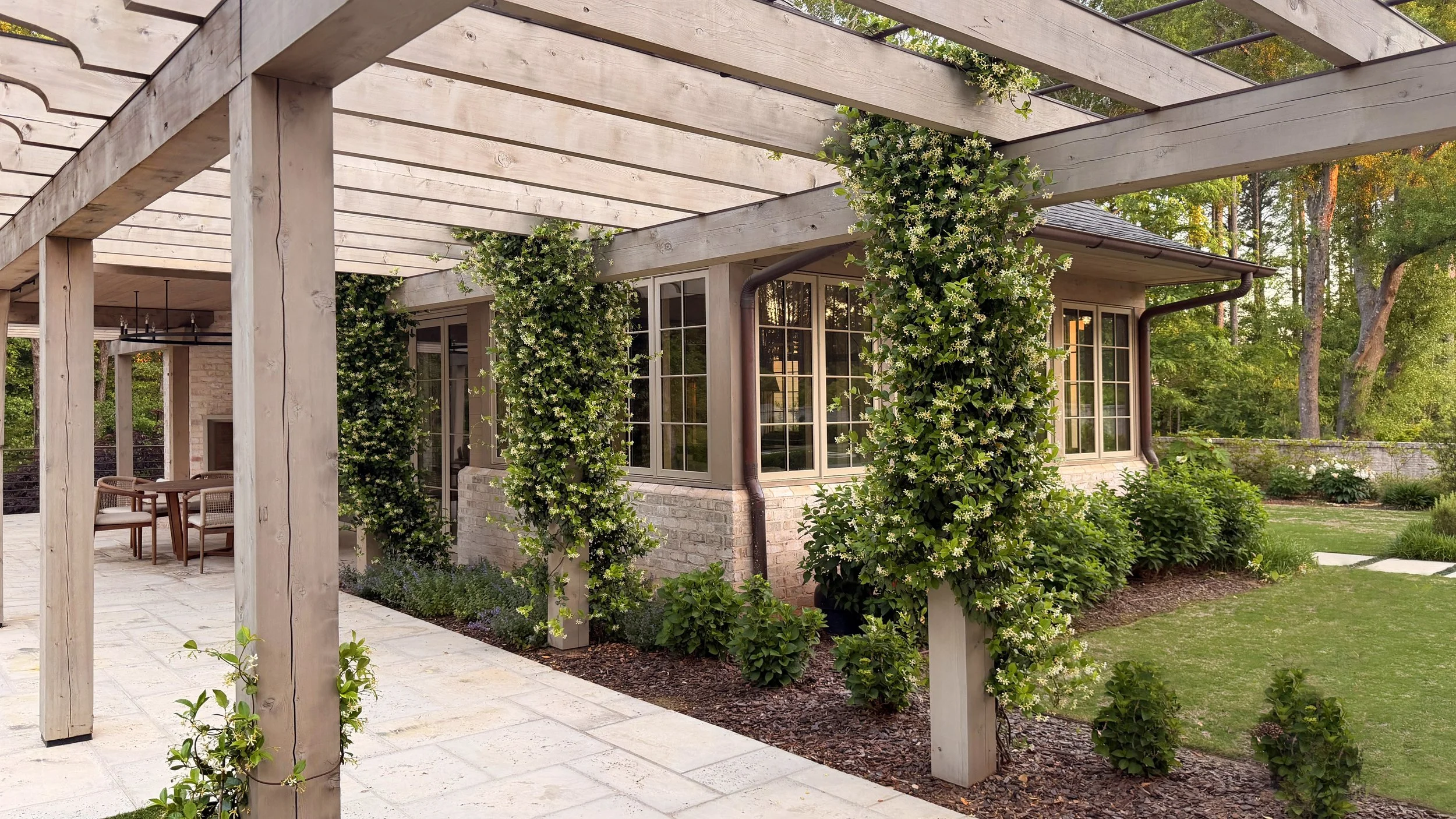 Exterior view of a house with large windows, surrounded by greenery and flowering vines, with a wooden pergola in the foreground and an outdoor seating area.