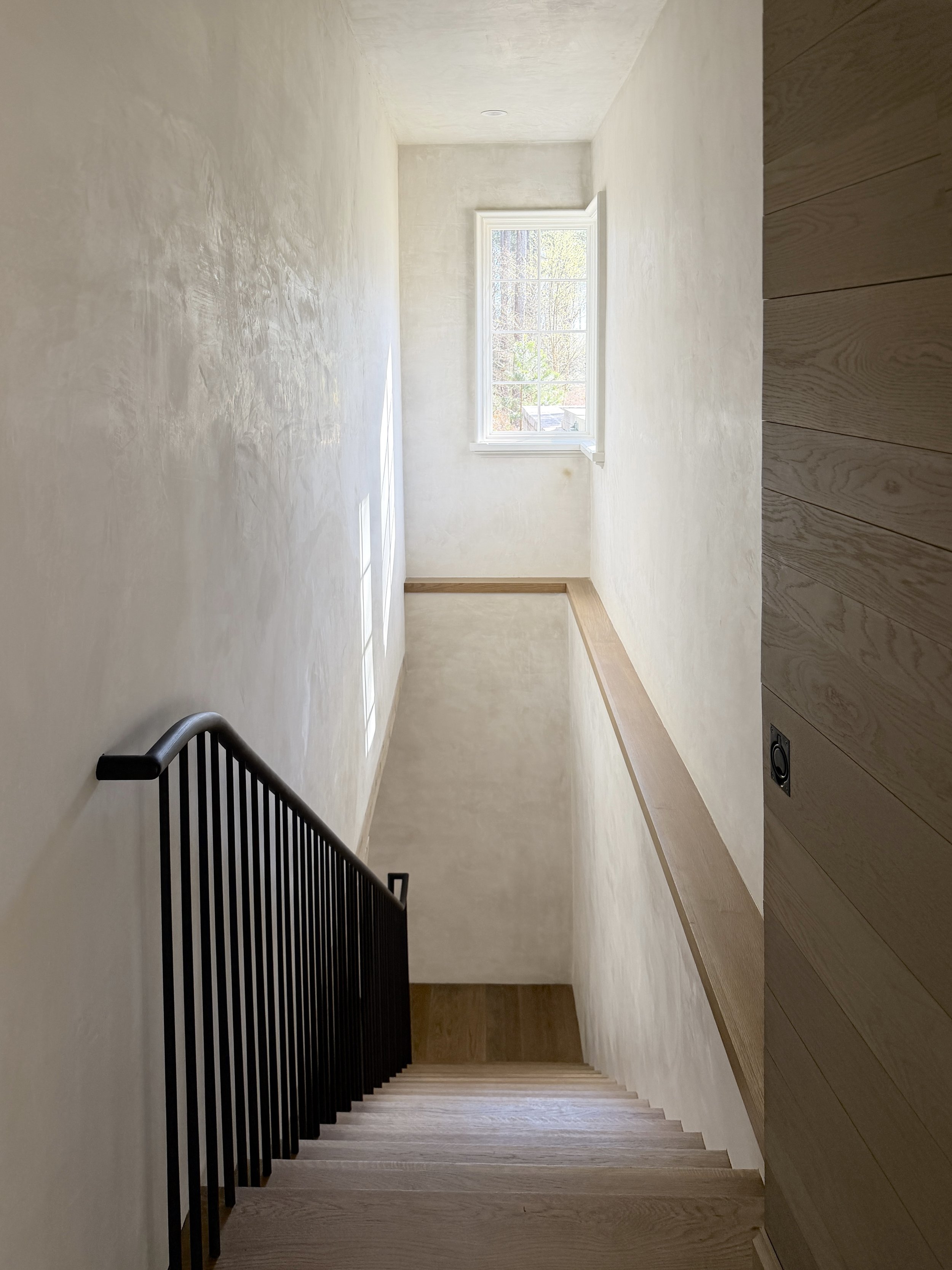 A staircase with wooden steps and a black metal railing on the left side, leading up to a small landing with a large window on the far wall, allowing natural light into the room.