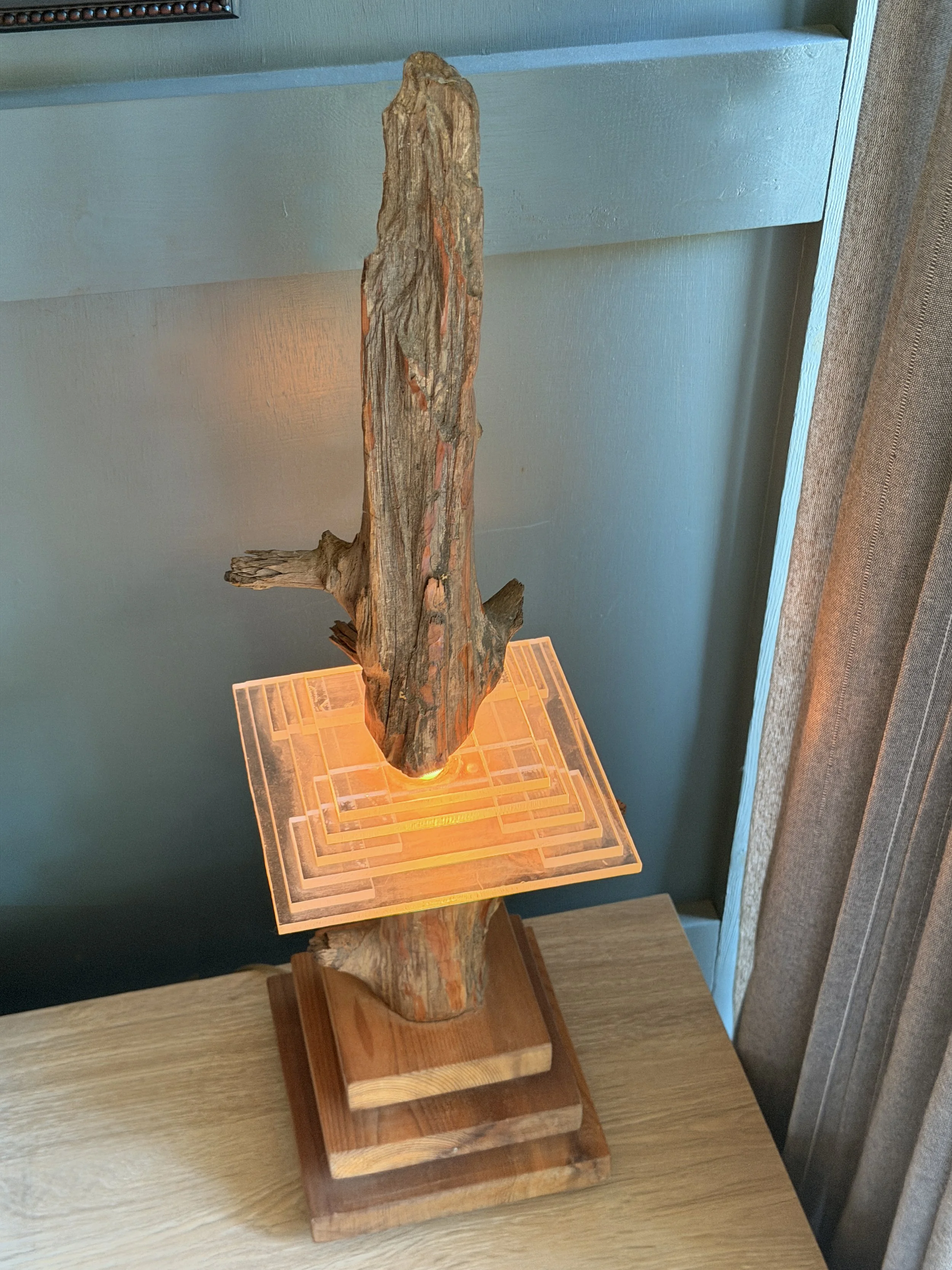 A carved wooden sculpture standing on a wooden pedestal, illuminated by warm lighting, against a light blue wall with a small display case nearby.