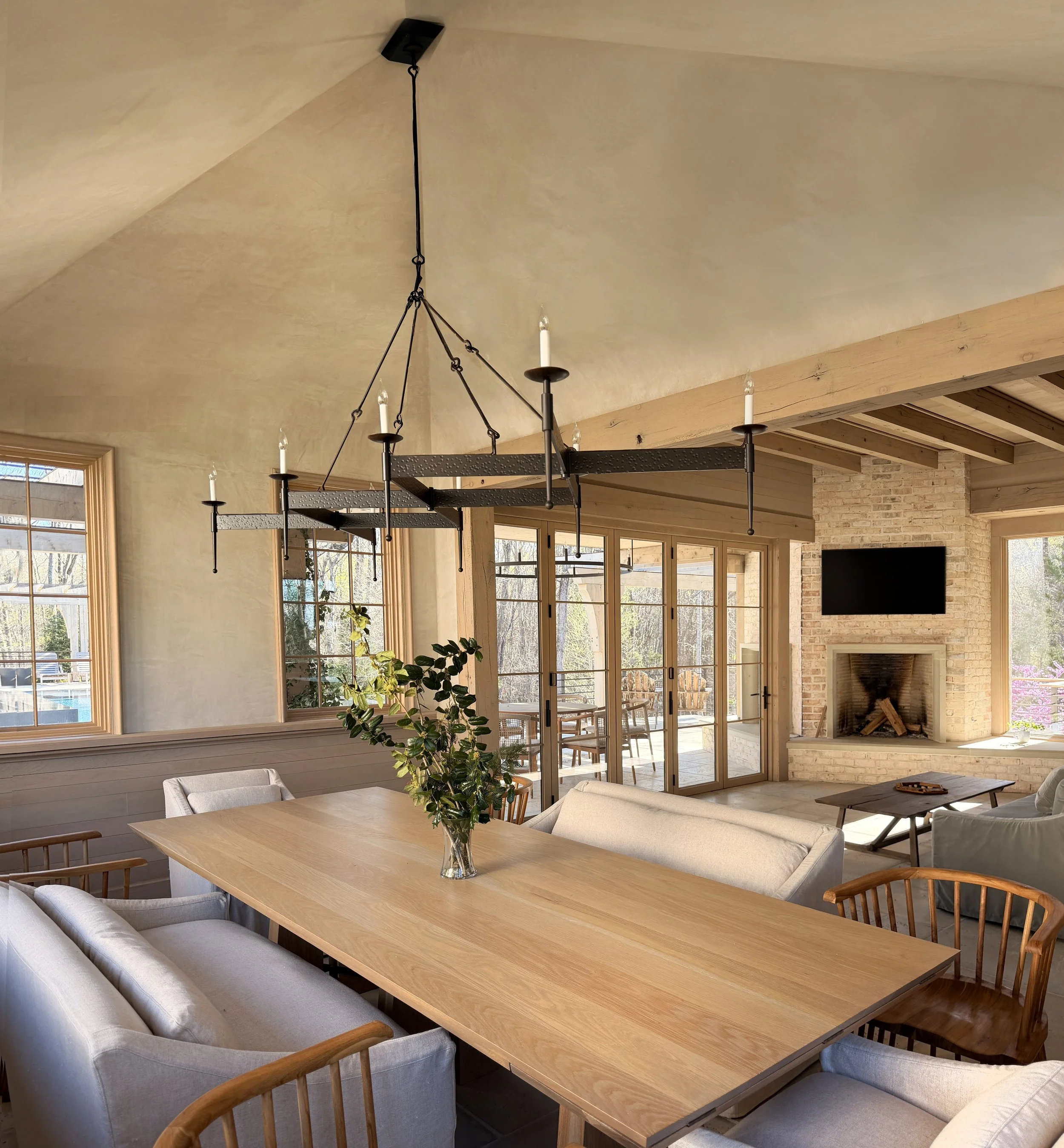 A dining area with a large wooden table, surrounded by mix of upholstered and wooden chairs, a vase with greenery, large windows with wooden frames, and a fireplace on a brick wall.