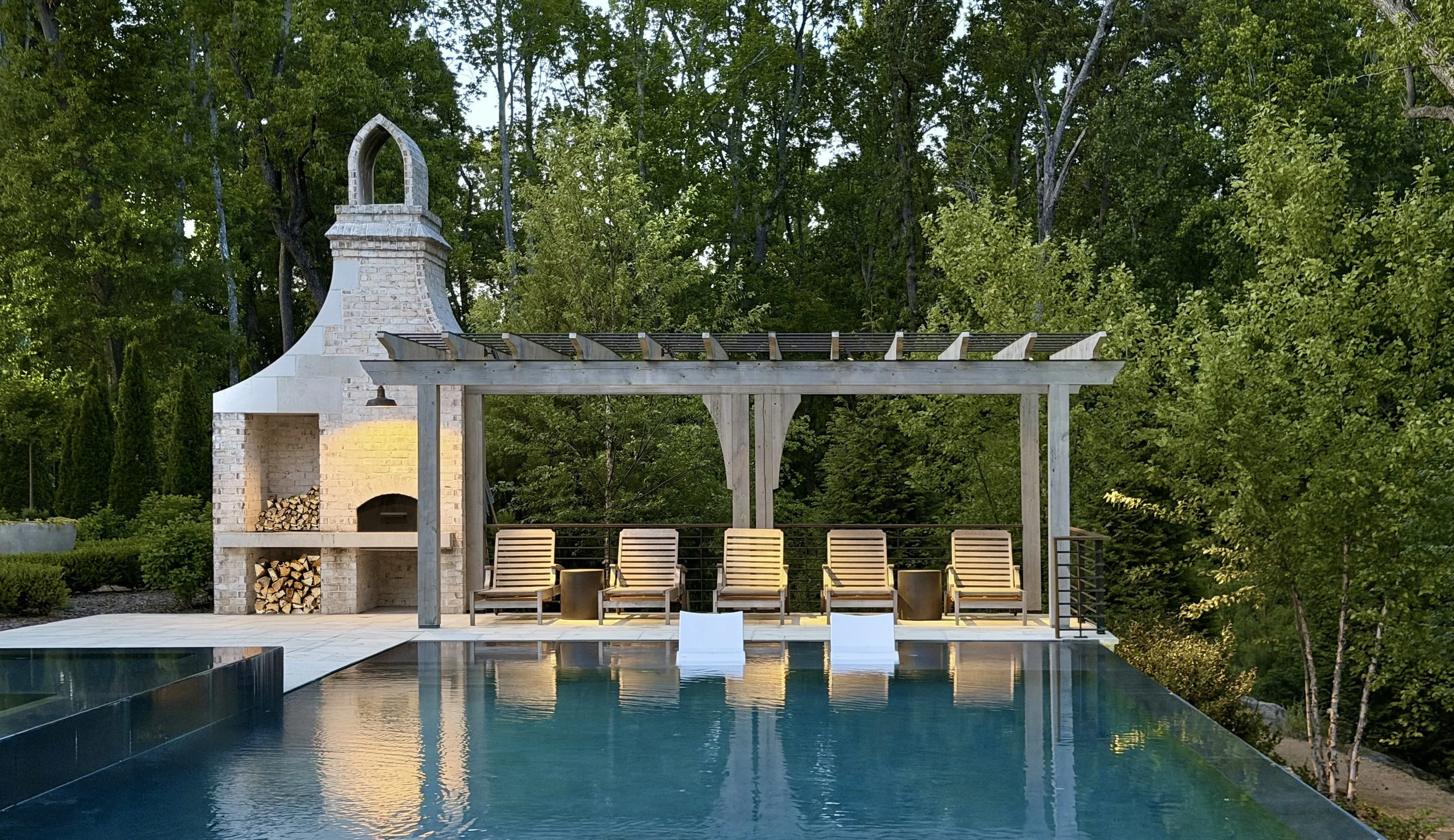 An outdoor patio area with a swimming pool in the foreground, a white brick outdoor fireplace and chimney, a wooden pergola, and a row of lounge chairs, surrounded by green trees.