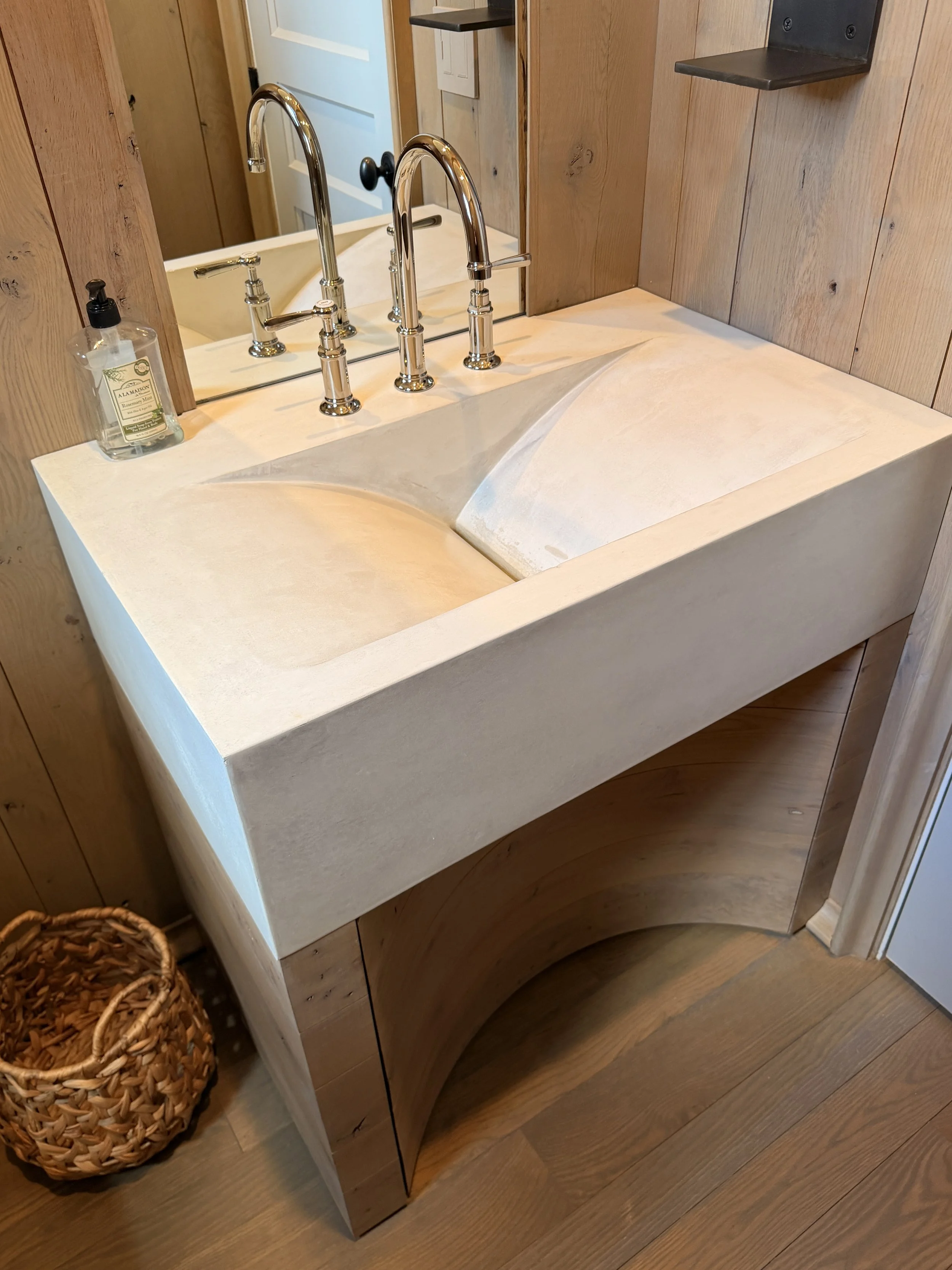 A bathroom vanity with a cream-colored countertop, a large mirror, and two shiny silver faucets. A bottle of hand soap sits on the left side of the sink. The surrounding walls are made of light wood, and there is a woven basket on the floor to the le