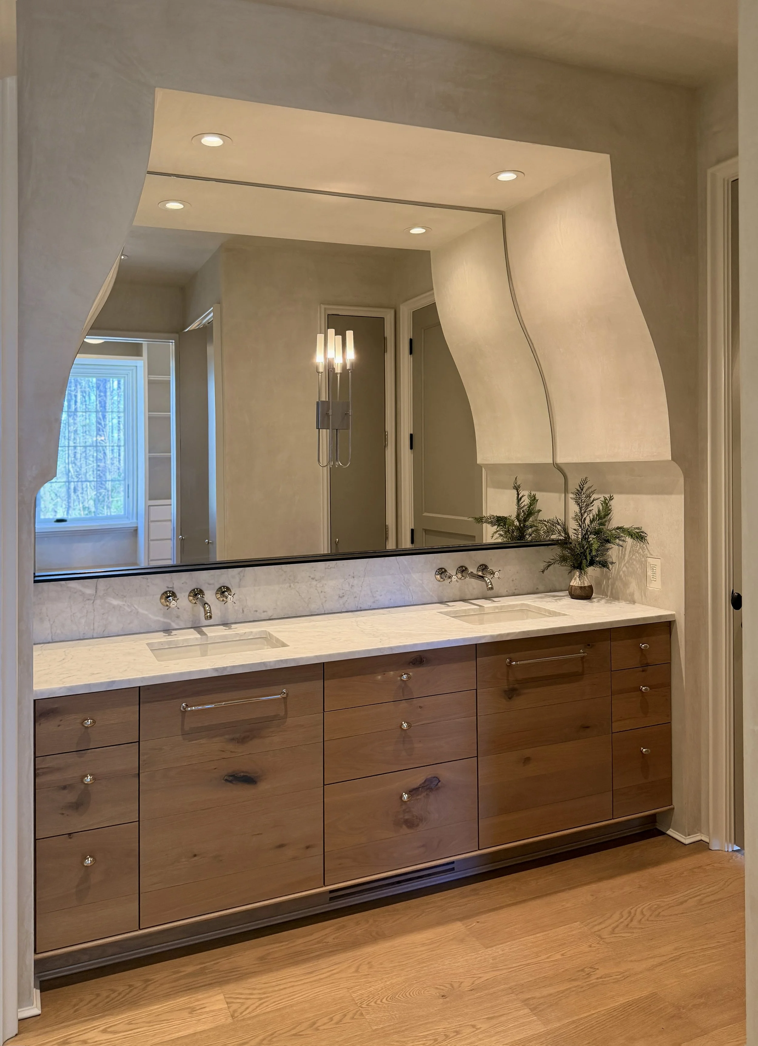 A bathroom vanity with a large mirror, wooden cabinets, marble countertop, and a vase with greenery. Recessed lighting and a window are visible in the reflection.