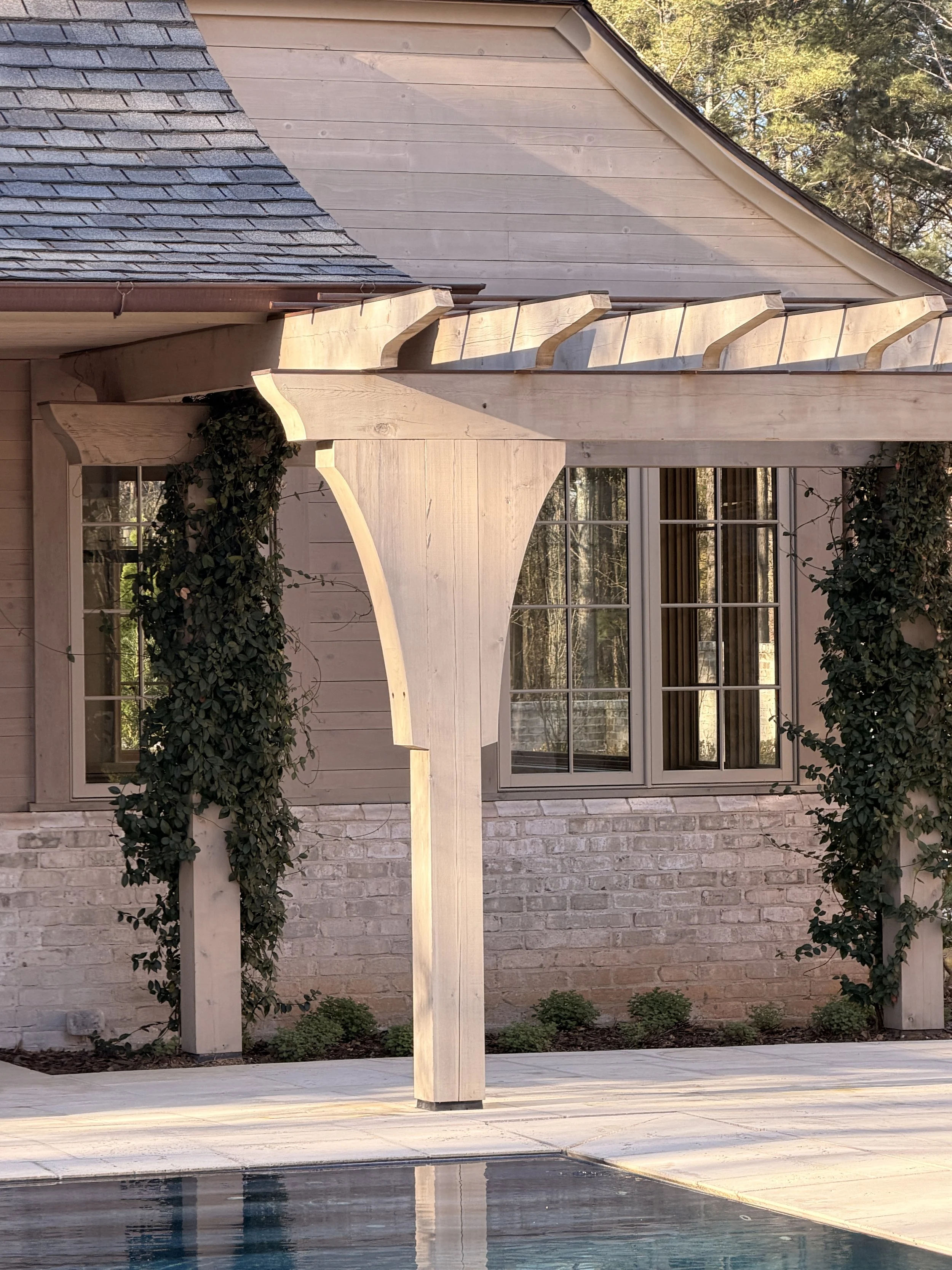 A backyard scene featuring a wooden pergola with decorative beams, a pool in the foreground, a brick house with large windows in the background, and trees with green foliage surrounding the area.