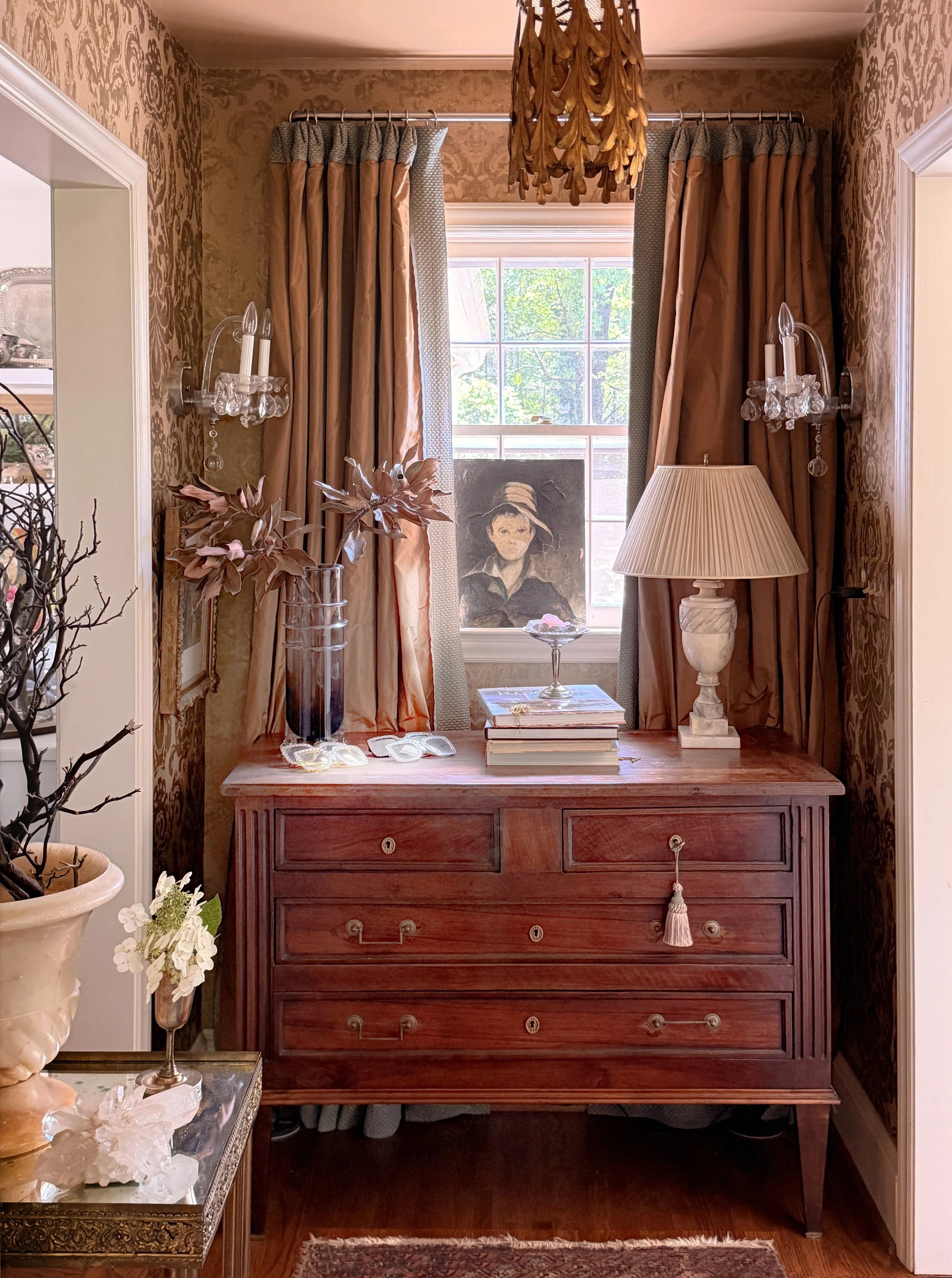 A wooden dresser with multiple drawers, topped with a stack of books, a glass bowl, and decorative objects. Behind it, a window with beige and pink curtains, and a portrait of a woman wearing a hat is visible on the windowsill. The room has walls wit