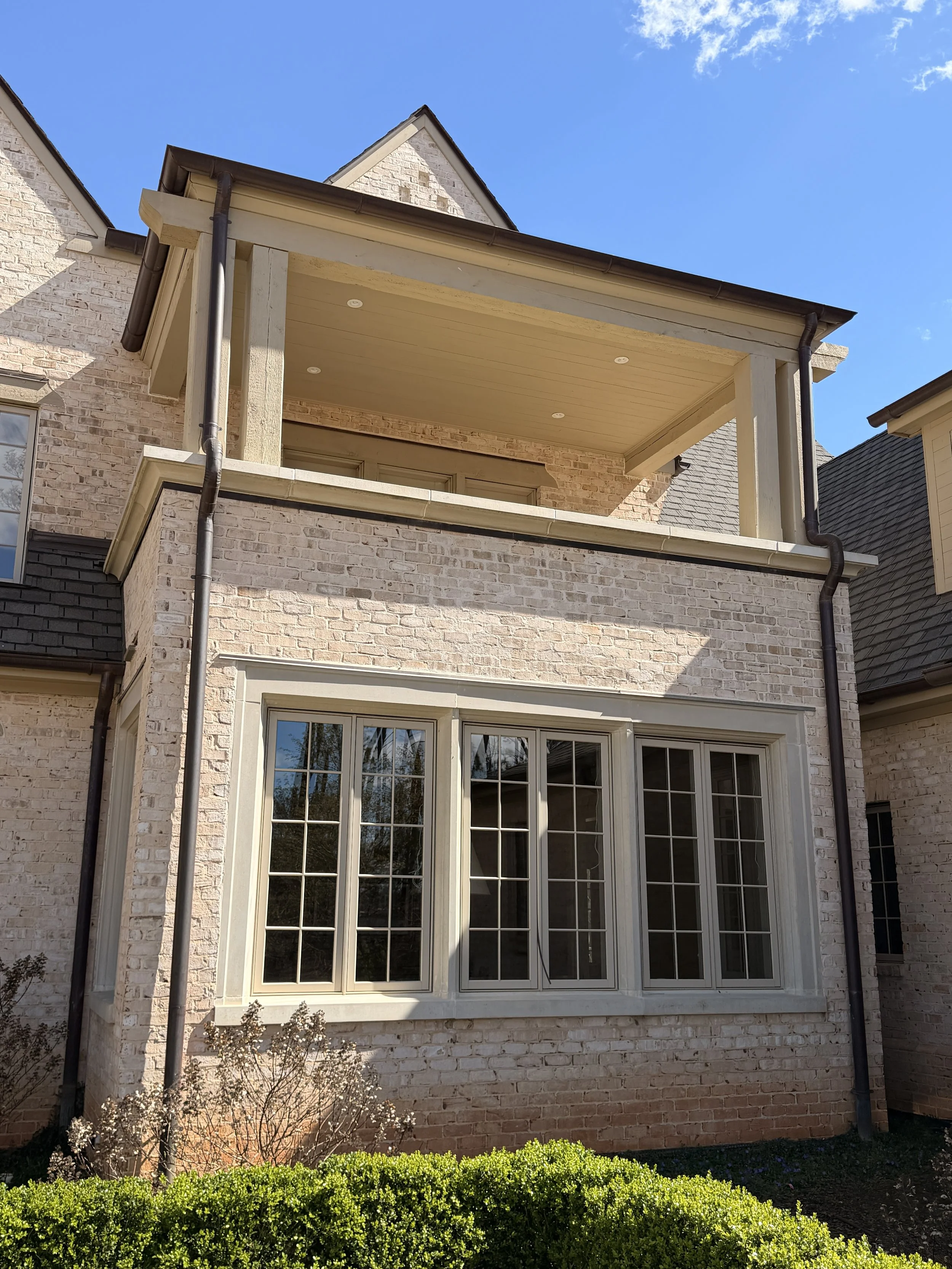 A two-story brick house with a large front window and a covered balcony on the second floor. The house has a sloped roof and rain gutters. There are bushes and plants in the yard in front of the house, and the sky is clear with a few clouds.