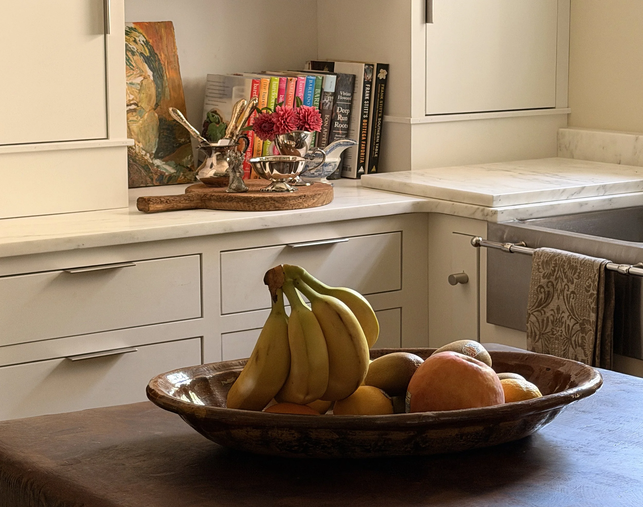 A wooden bowl filled with bananas, peaches, and other fruit on a kitchen counter, with books, a painting, and flowers in the background.