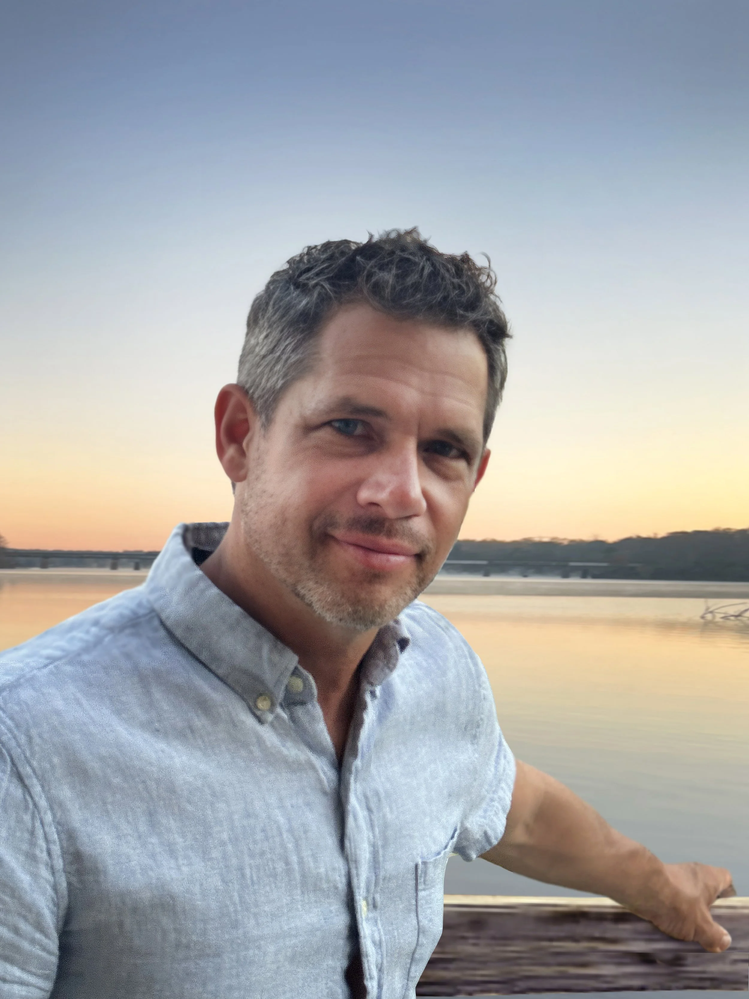 A man with curly gray hair and blue eyes wearing a light blue button-up shirt, standing on a bridge by a river during sunset, with his arm resting on the railing and looking at the camera.