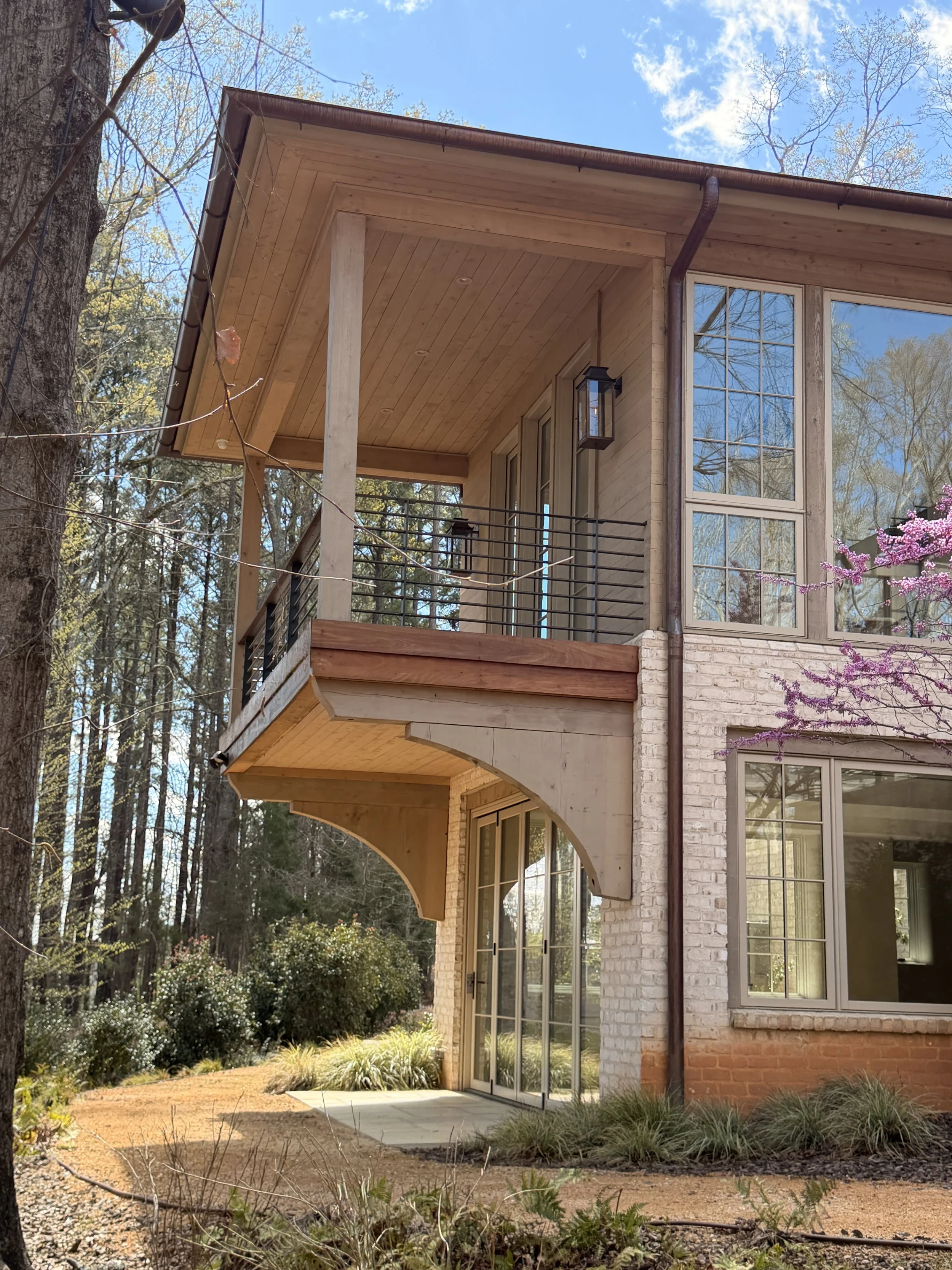 Back porch of a house with large windows, wooden ceiling, and outdoor lighting, surrounded by trees and shrubs.