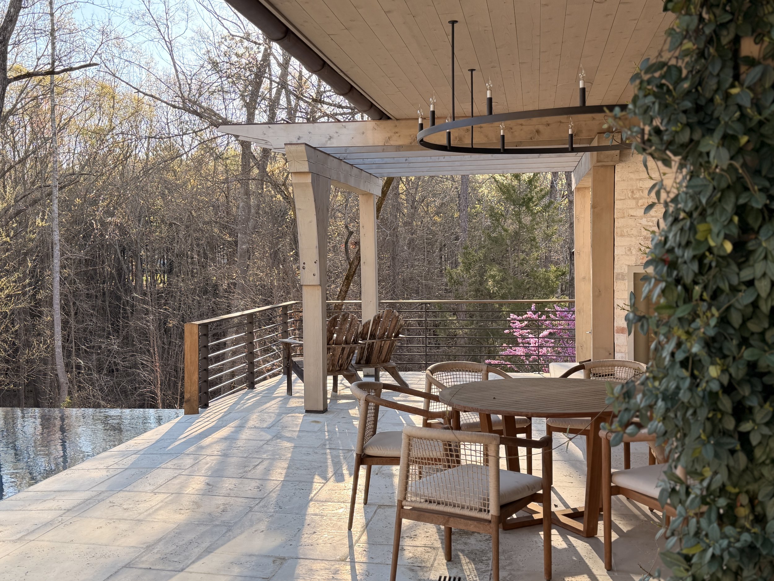 A patio area with patio furniture including chairs around a table, overlooking a wooded backyard with trees and a flowering shrub, on a sunny day.