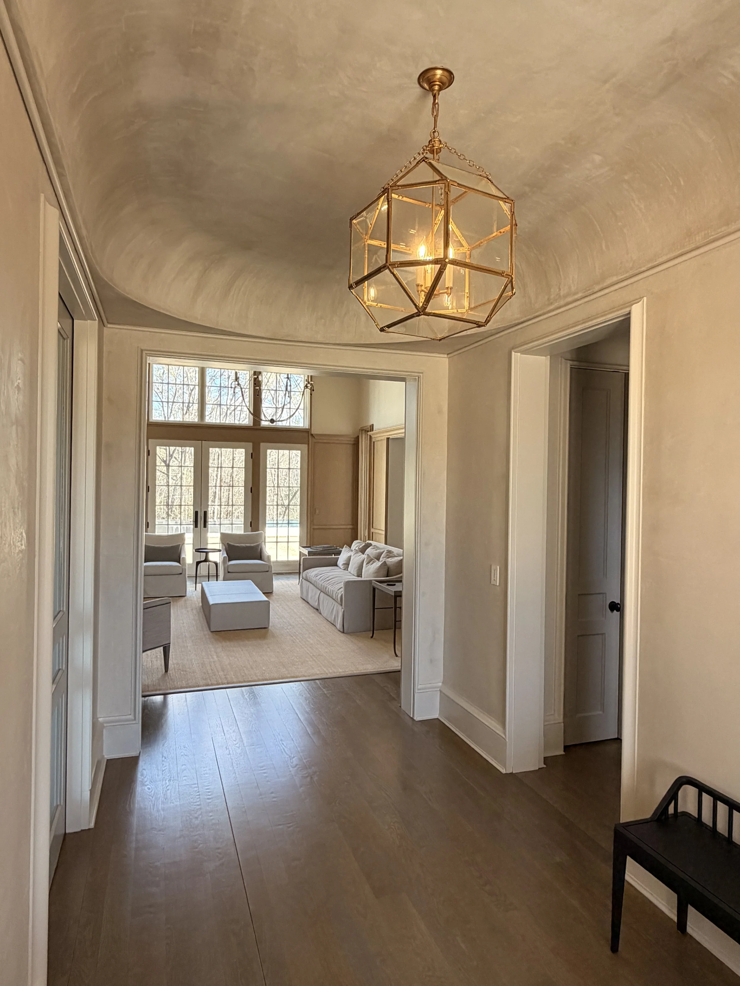 A spacious living room with large windows, cream-colored walls, and hardwood flooring. There are two armchairs and a sofa with cushions, a small table, and a woven rug. The room is lit by sunlight and a pendant light hanging from the ceiling.